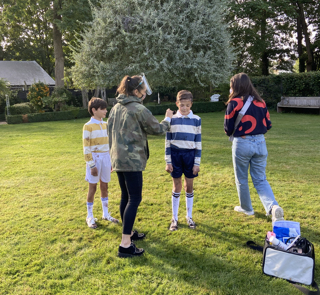 Two young boys in sports uniforms and two adult women stand on a lush green field, with one woman applying face paint to a boy's face, during what appears to be a sporting event or outdoor activity.