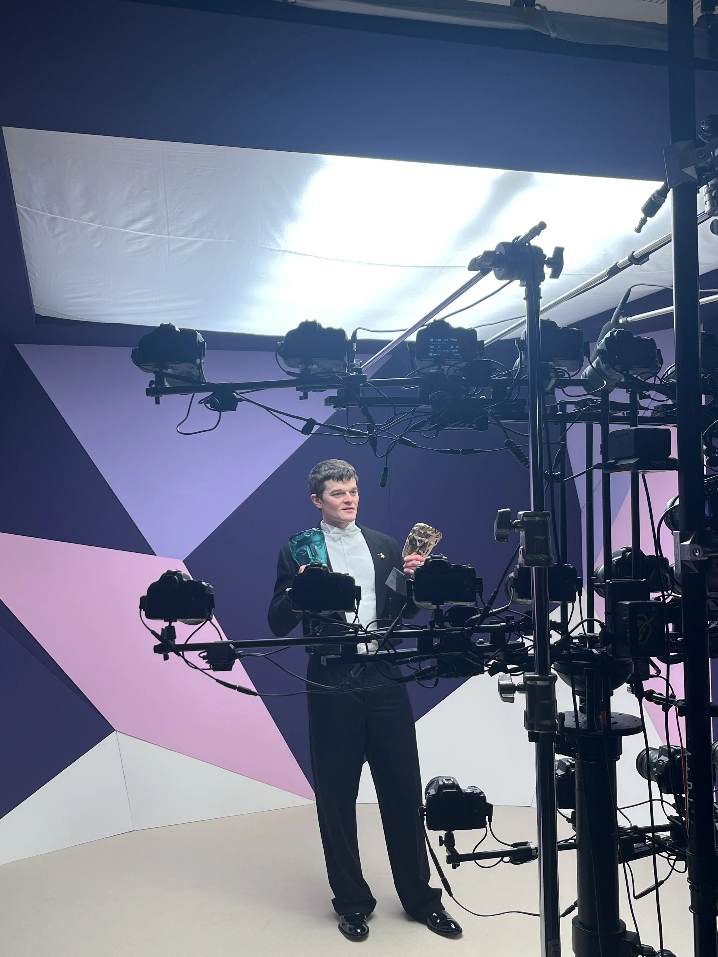 A man in formal attire, holding two BAFTA awards appears to be in a professional photo or video shoot studio with lighting equipment and a geometric, colorful backdrop.