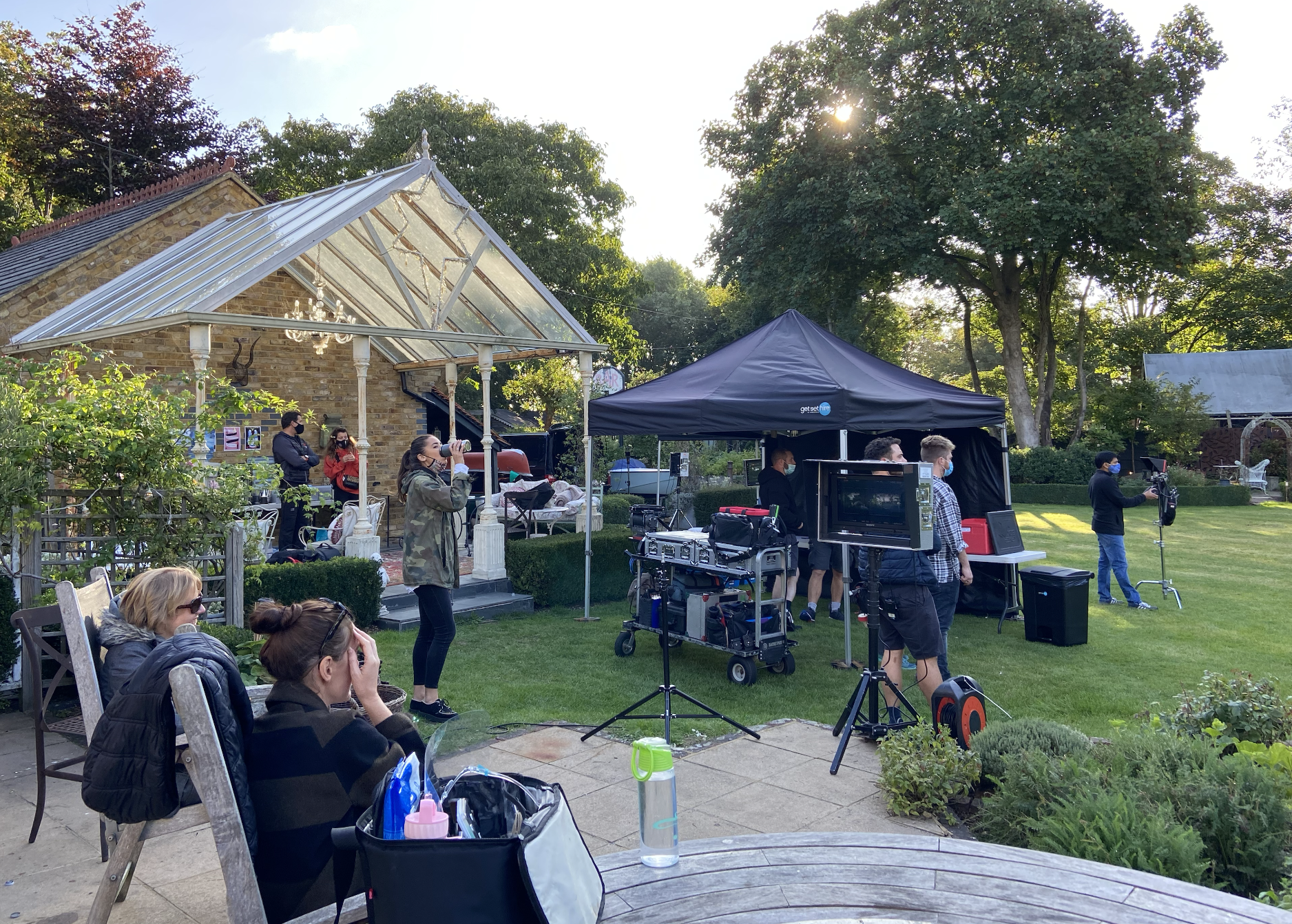 Filming setup in a backyard garden with crew members, equipment, and a small audience.