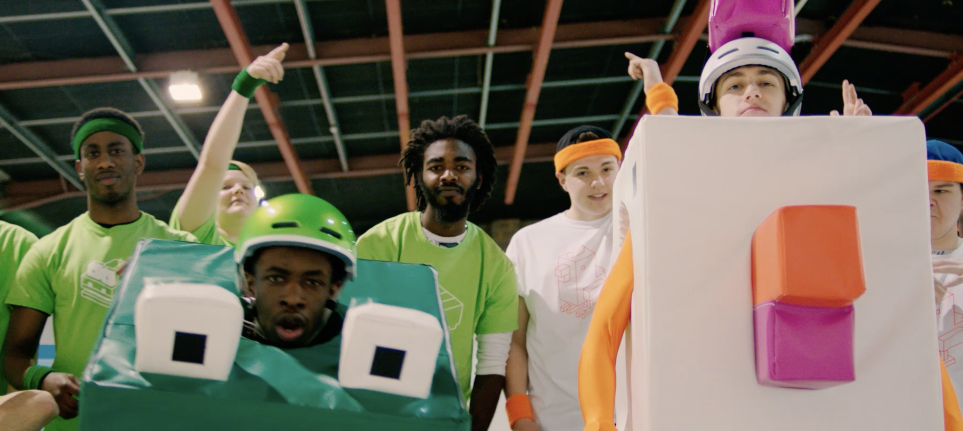 Group of people dressed in costumes resembling Tetris game characters, standing inside a gymnasium or warehouse with a high ceiling. The performers are wearing bright outfits and helmets, with some making expressive gestures or poses.