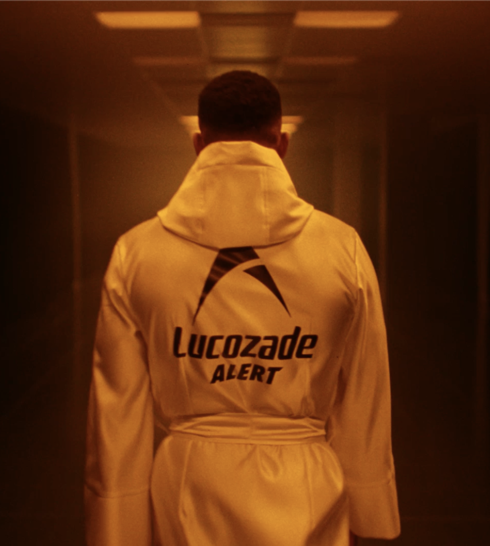 A person in a yellow jacket with a hood, walking down a dimly lit corridor. The back of the jacket has a logo and the words 'Lucozade ALERT'.