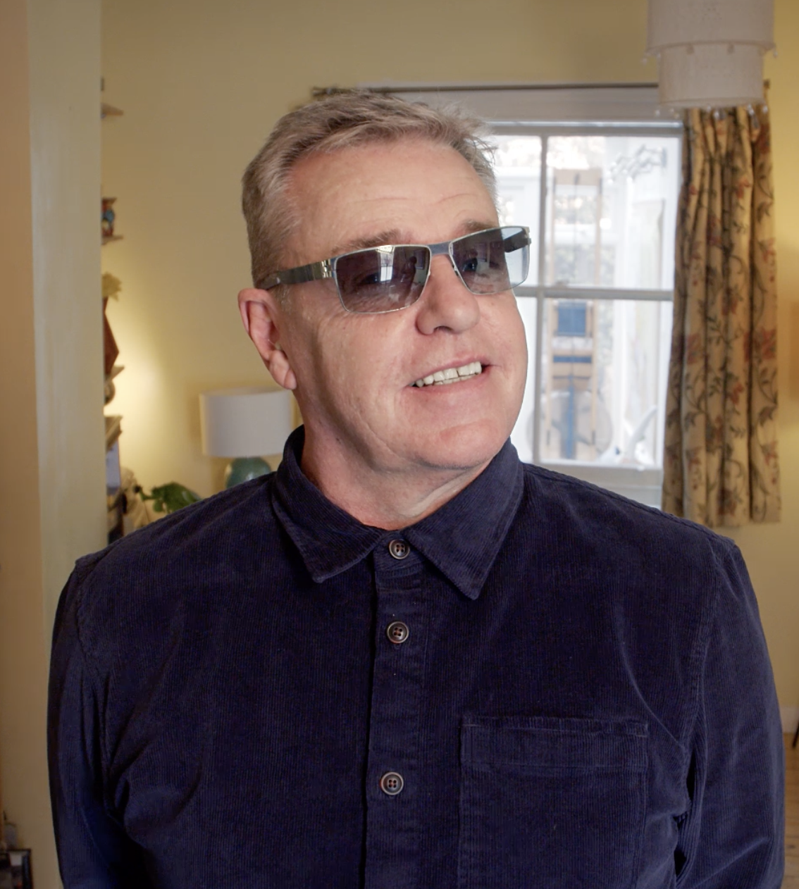Suggs wearing sunglasses and a dark button-up shirt inside a home with a window, beige walls, curtains, a table lamp, and decorative items in the background.