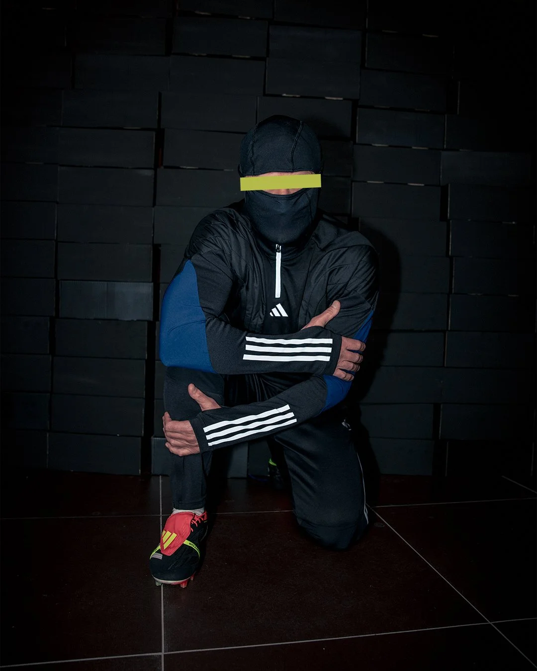 A person dressed in black athletic clothing with blue accents and Adidas stripes, wearing a face mask with a yellow reflective band over the eyes, is kneeling on one knee in front of a black wall, adjusting their shoe.