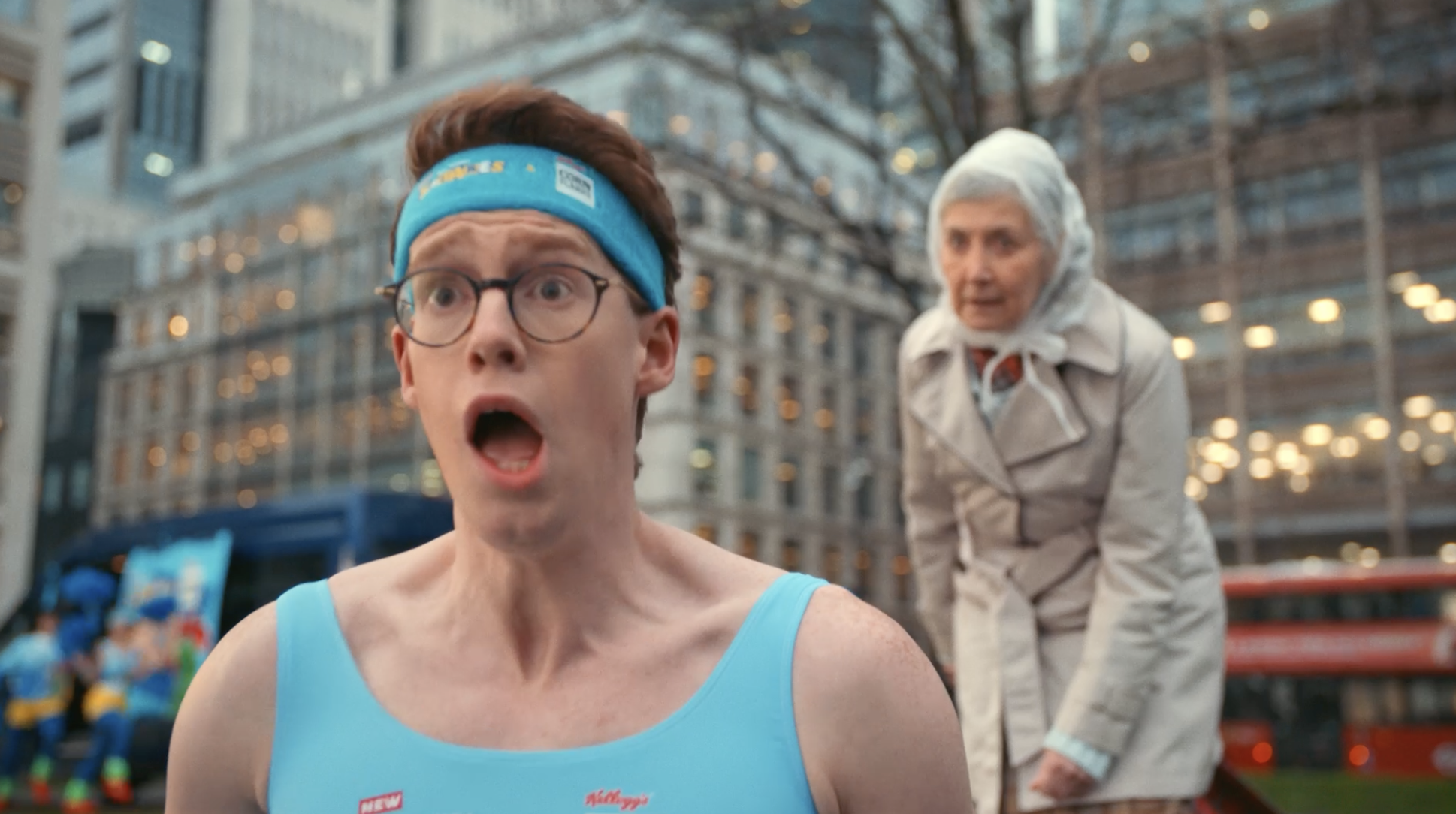 A young man wearing glasses and a blue headband, dressed in a blue athletic tank top, appears surprised with his mouth open. An older woman in a beige trench coat and a hairnet stands behind him in an outdoor urban setting.