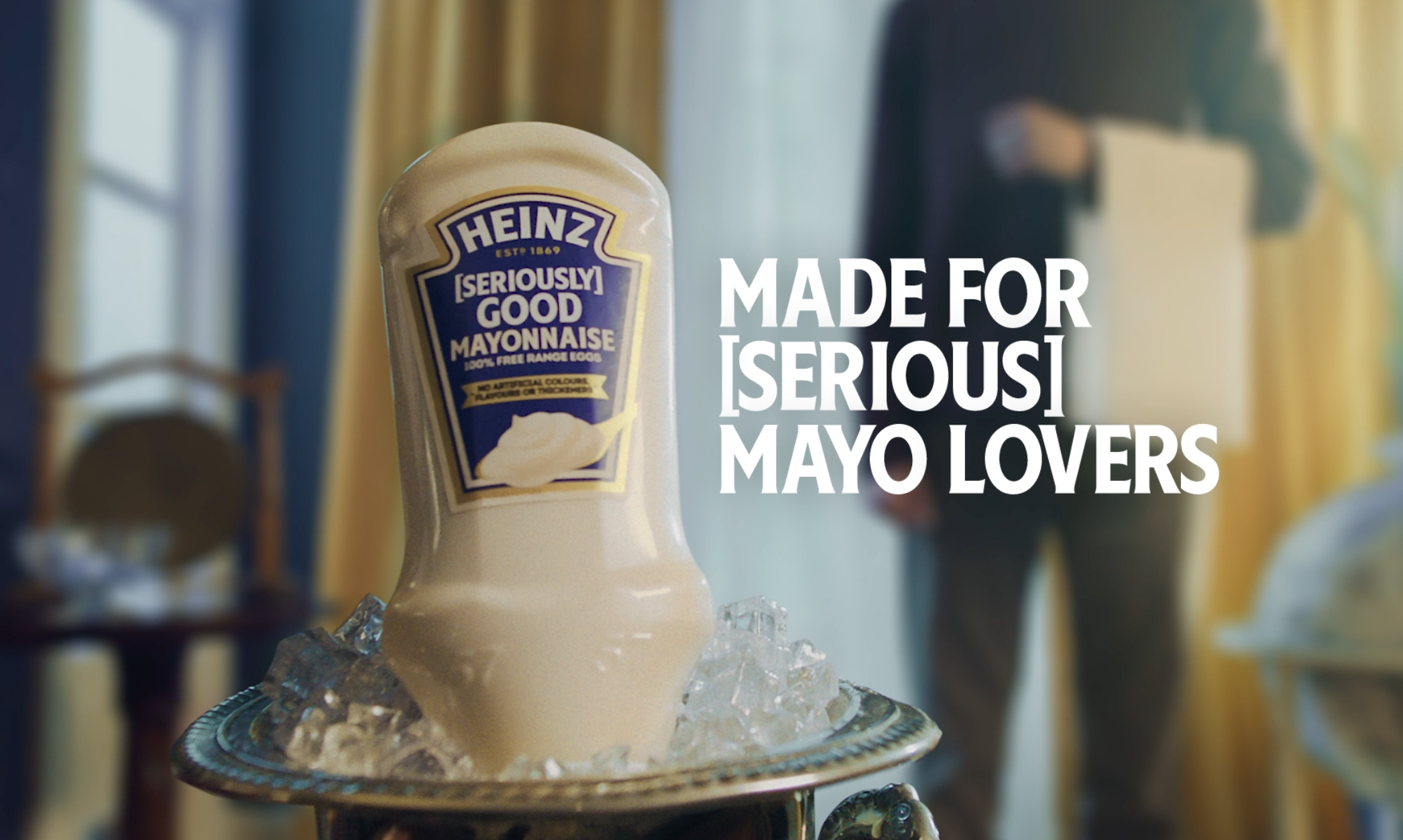 A bottle of Heinz Seriously Good Mayonnaise on a tray with ice, with text overlay that reads 'Made for [Seriously] Mayo Lovers' in a cozy room with yellow and blue curtains.