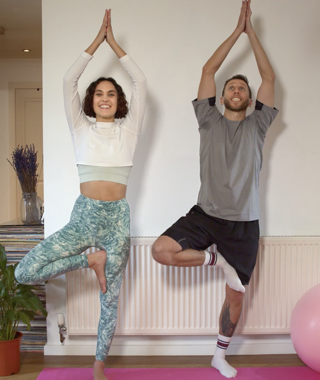A woman and a man practicing yoga indoors, standing on one leg in tree pose with hands together above their heads, smiling.