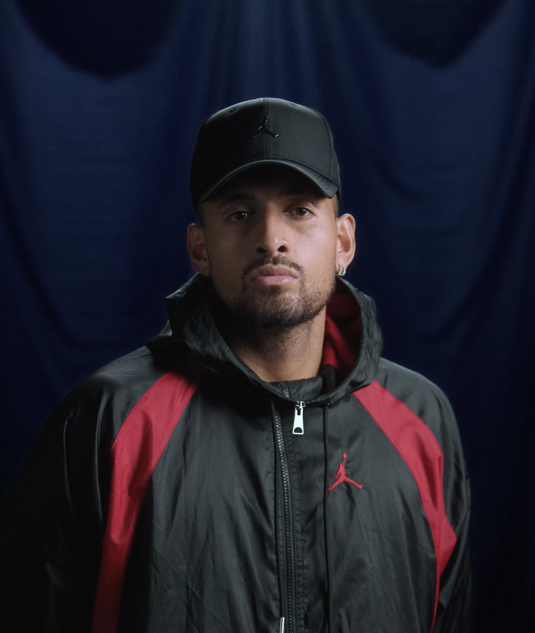Nick Kyrgios wearing a black cap and black and red athletic jacket with the Jordan logo, standing against a dark backdrop.