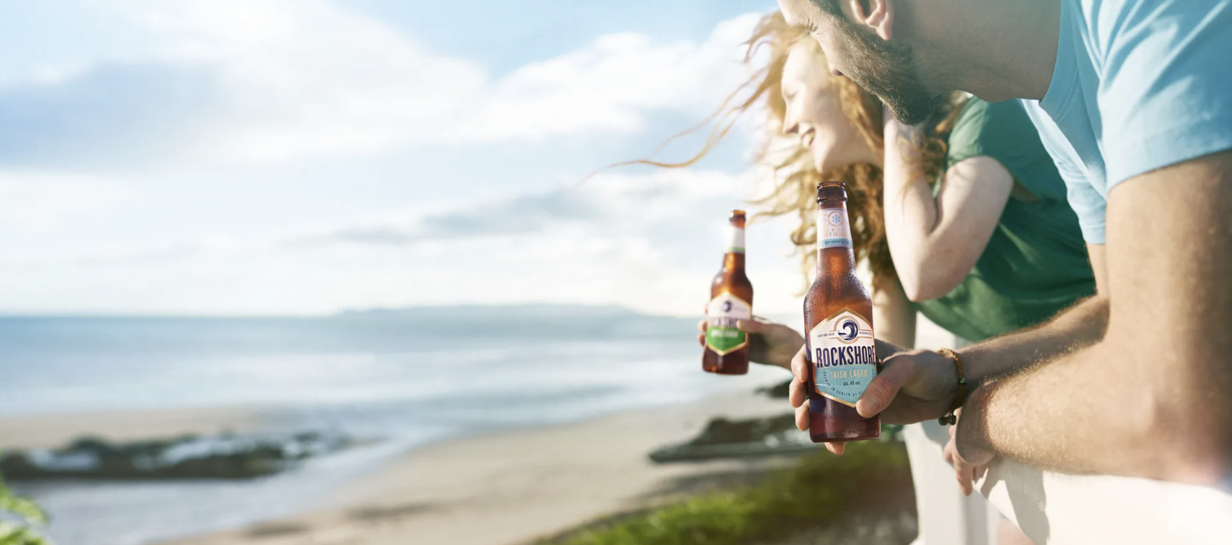 People holding beer bottles on a beach with ocean and sky in the background.