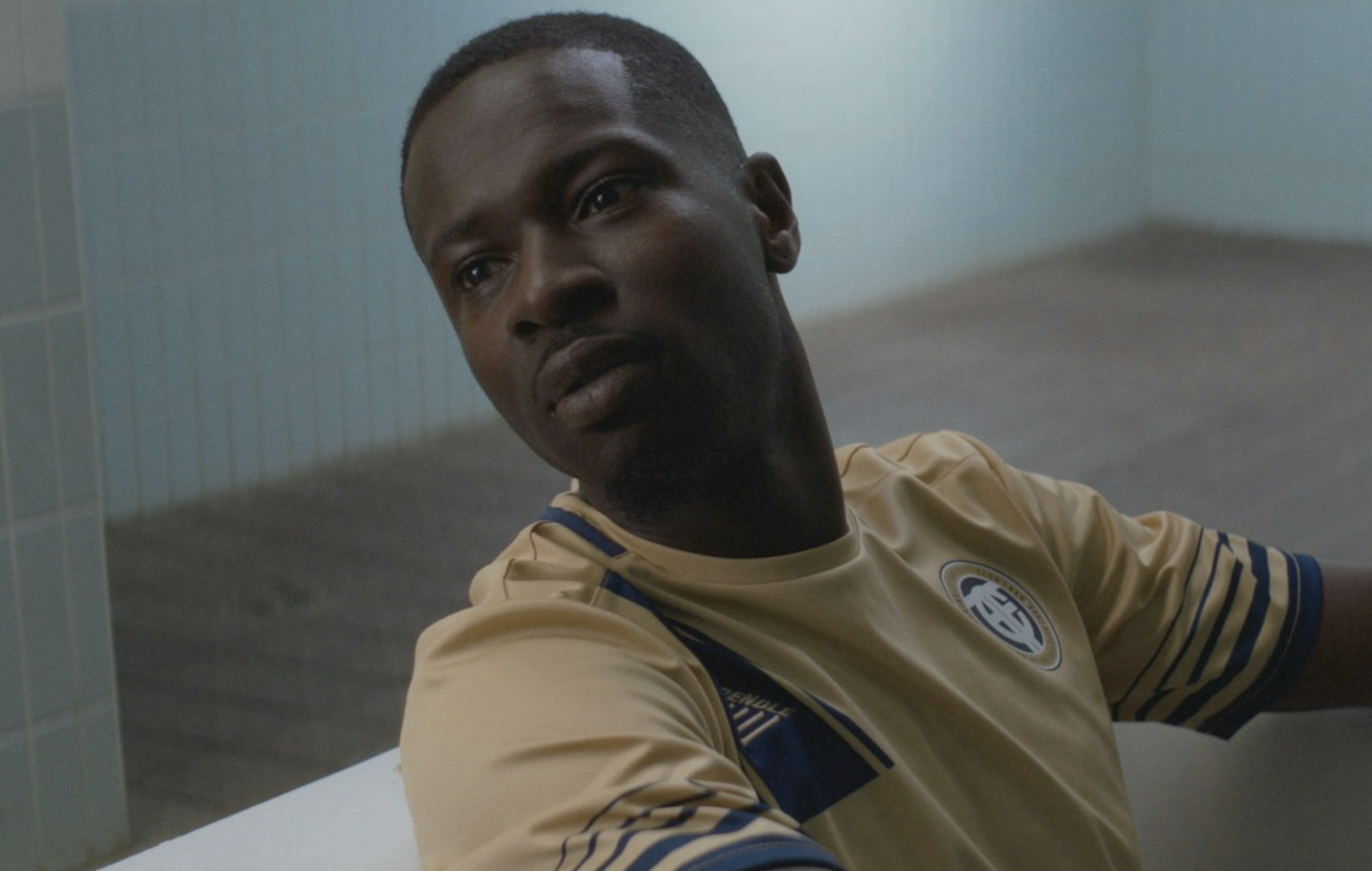 A young man with short hair sitting on a bed in a room, wearing a beige sports jersey with a logo on the chest.