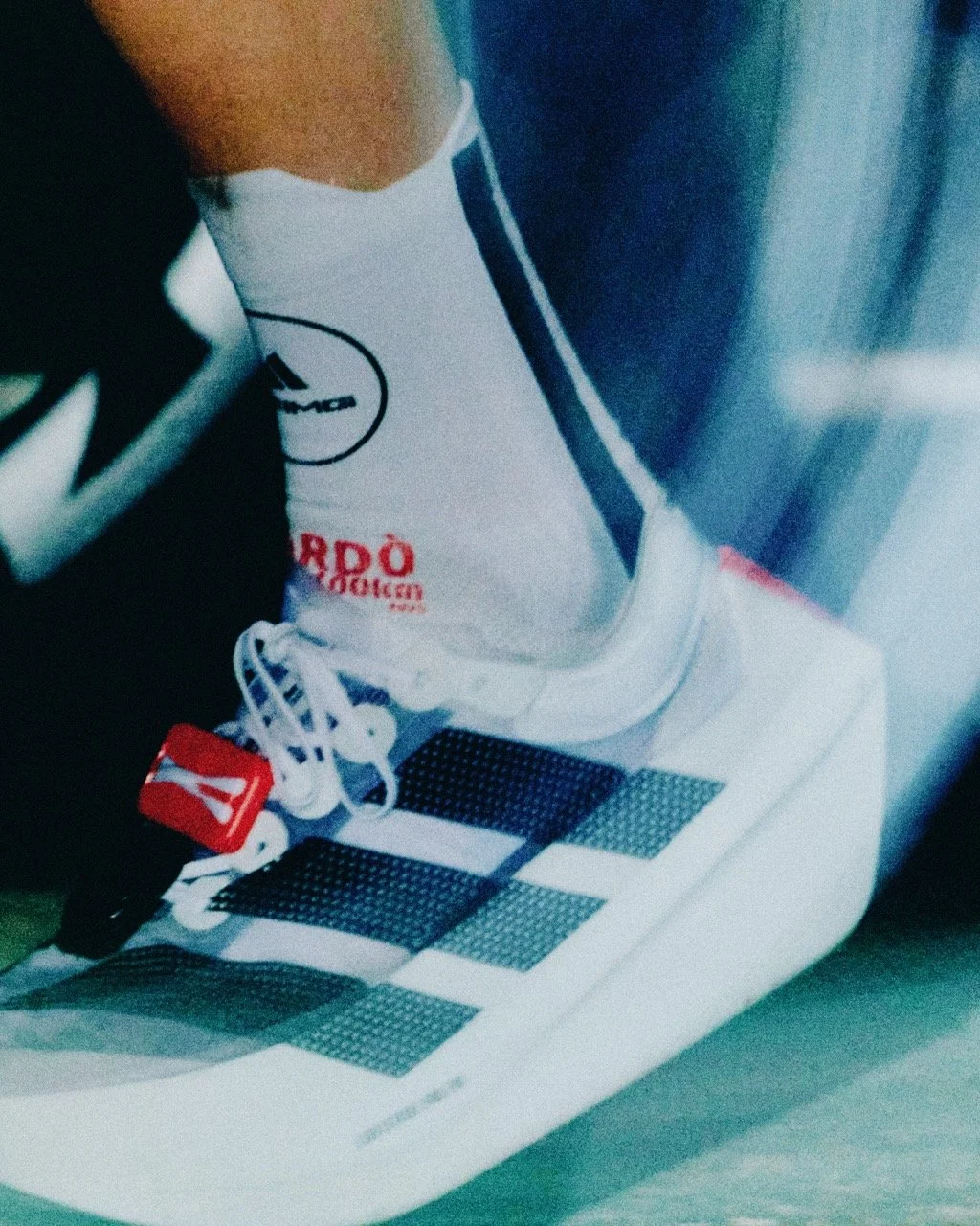 Close-up of a person's foot wearing a white Adidas sneaker with black and red accents, resting on a white bench with black and gray patches.