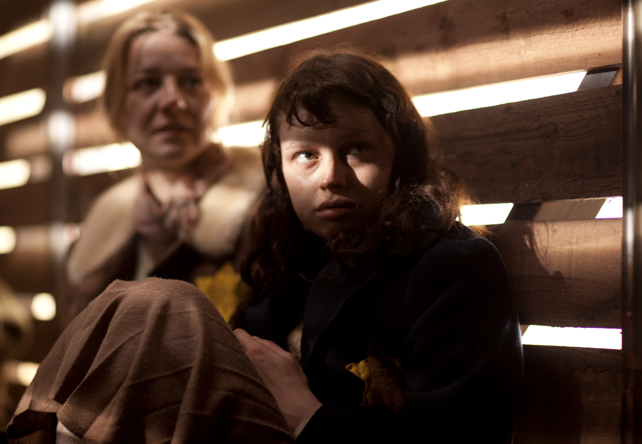 A young woman with curly hair sitting on a bench with a distressed expression, illuminated by slats of light, with an older woman blurred in the background, also sitting on the bench.