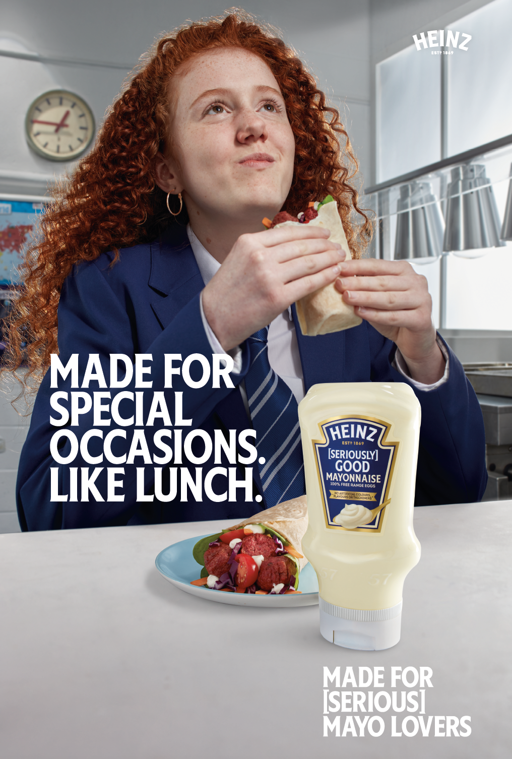 A girl with curly red hair in a blue school uniform eating a wrap sandwich in a kitchen or cafeteria. There is a plate of colorful salad and a bottle of Heinz mayonnaise on the table. Text on the image says, 'Made for special occasions. Like lunch.' 