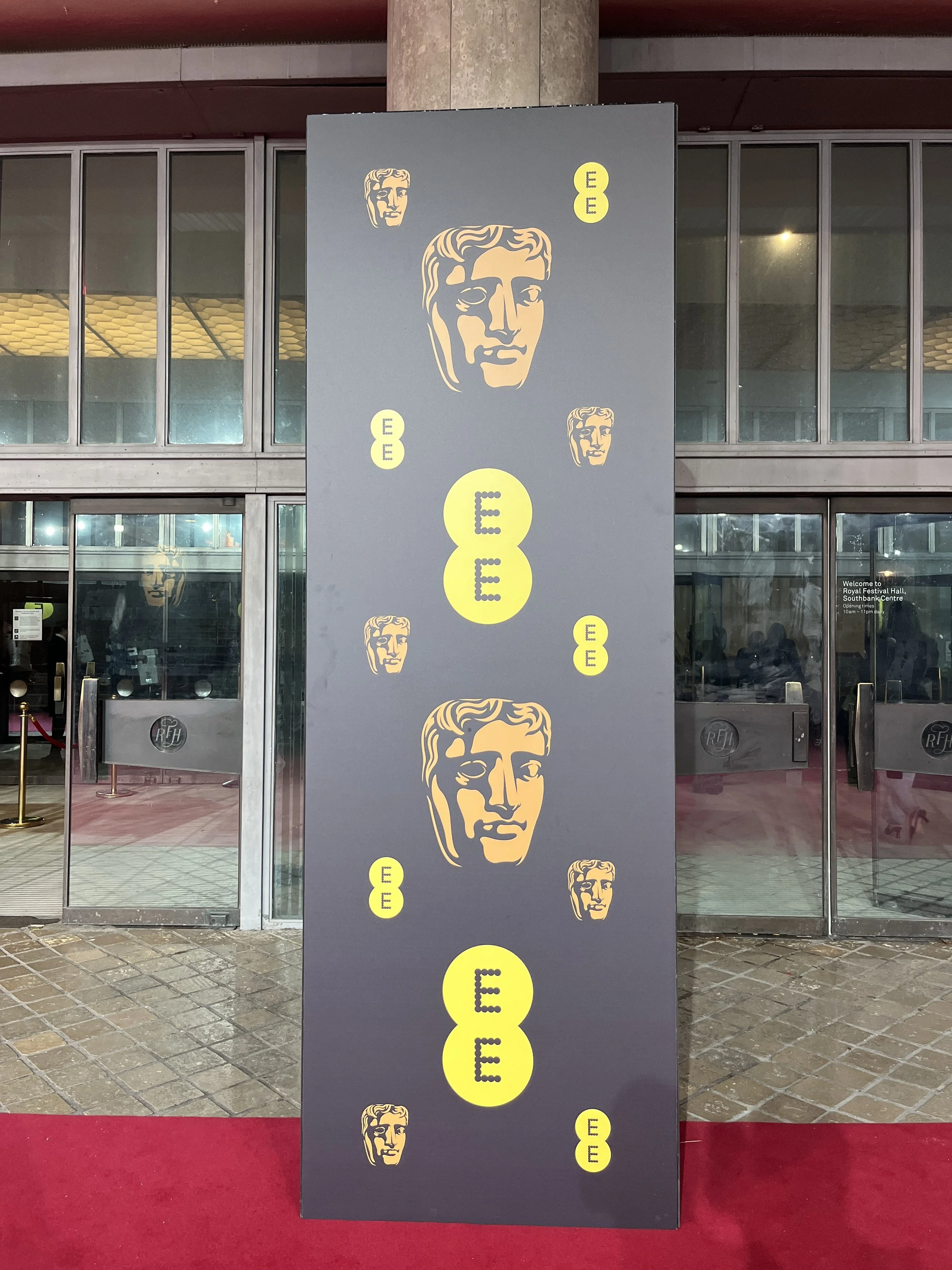 A vertical rectangular sign with a dark background featuring multiple yellow circles and stylized headshot illustrations of a man, all displaying the letter 'E'. The sign is placed outside a building with glass doors and windows, with a red carpet in front.