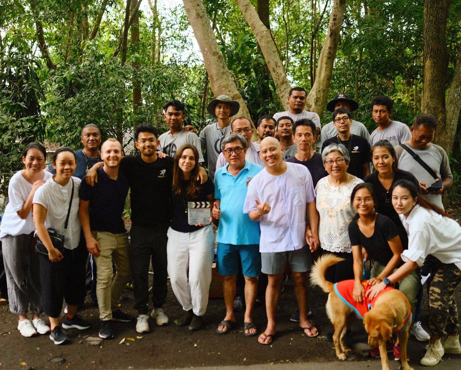 Group of diverse people posing outdoors in a wooded area, with one person holding a film slate and a woman with a dog in a costume.