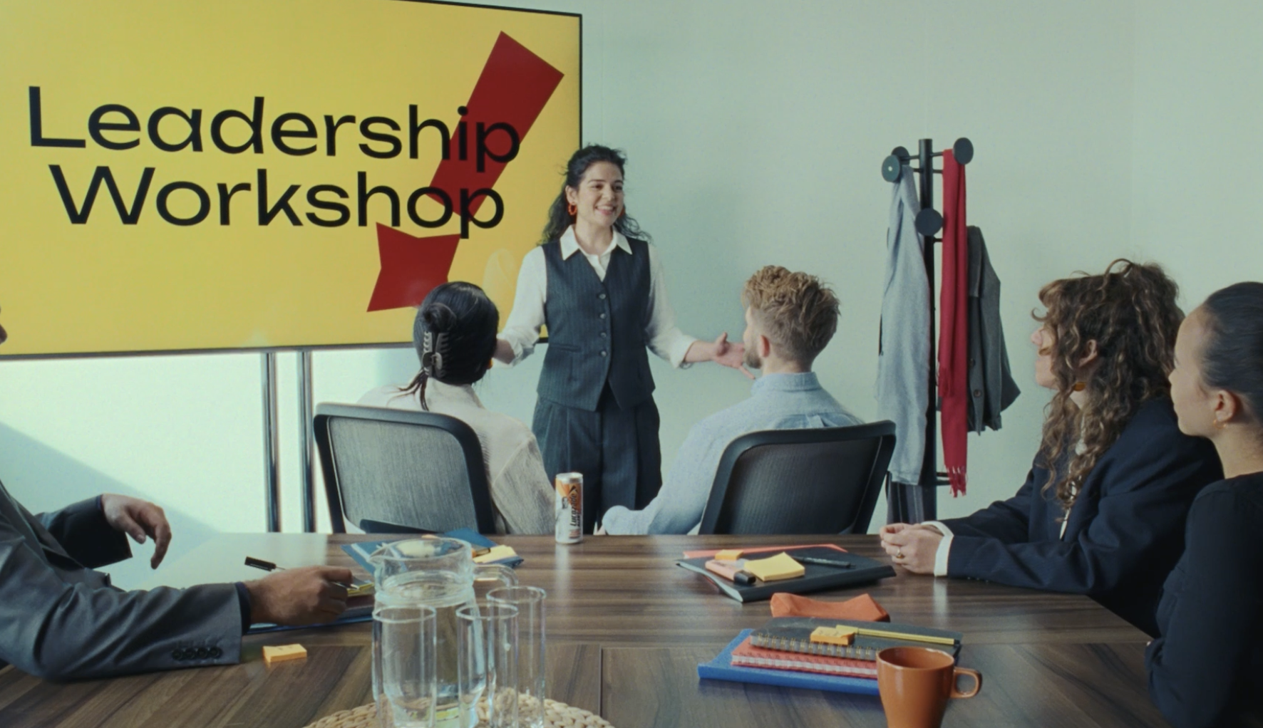 A woman leading a leadership workshop in a conference room with four seated participants, a large screen displaying 'Leadership Workshop,' and office supplies on the table.