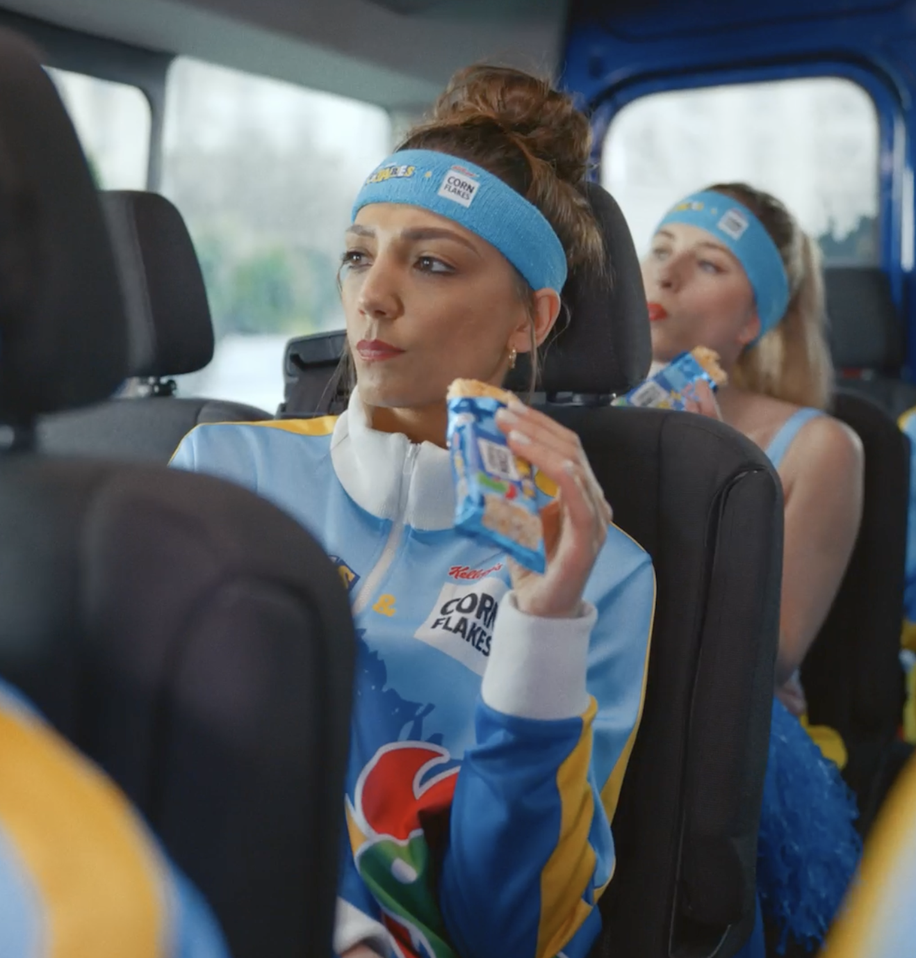 Two women wearing matching blue headbands and sports uniforms sitting in a vehicle and eating snack bars.