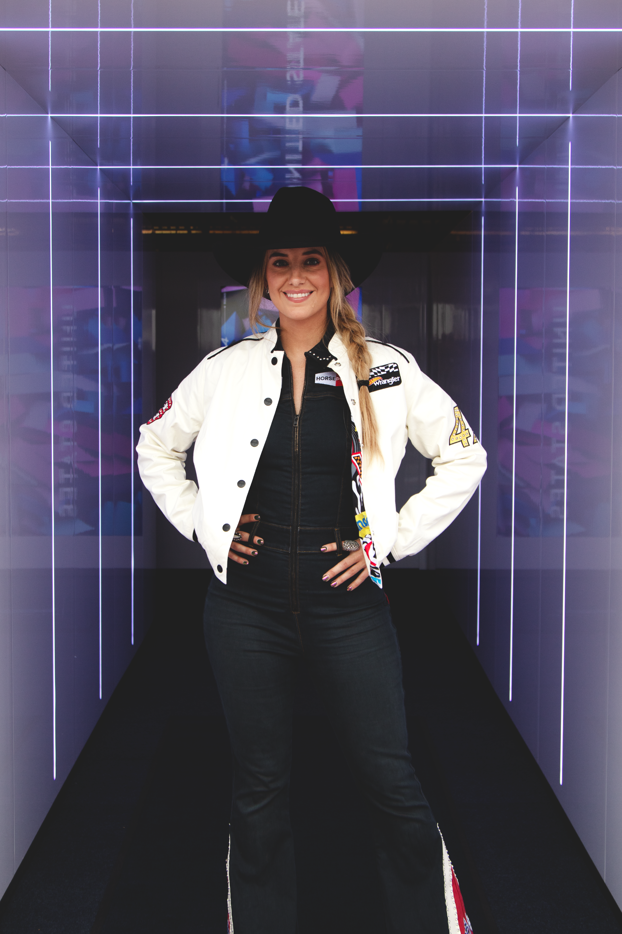 A woman in racing attire, including a white jacket with sponsor patches, a black shirt, and a wide-brimmed black hat, standing in a tunnel with purple LED lights.