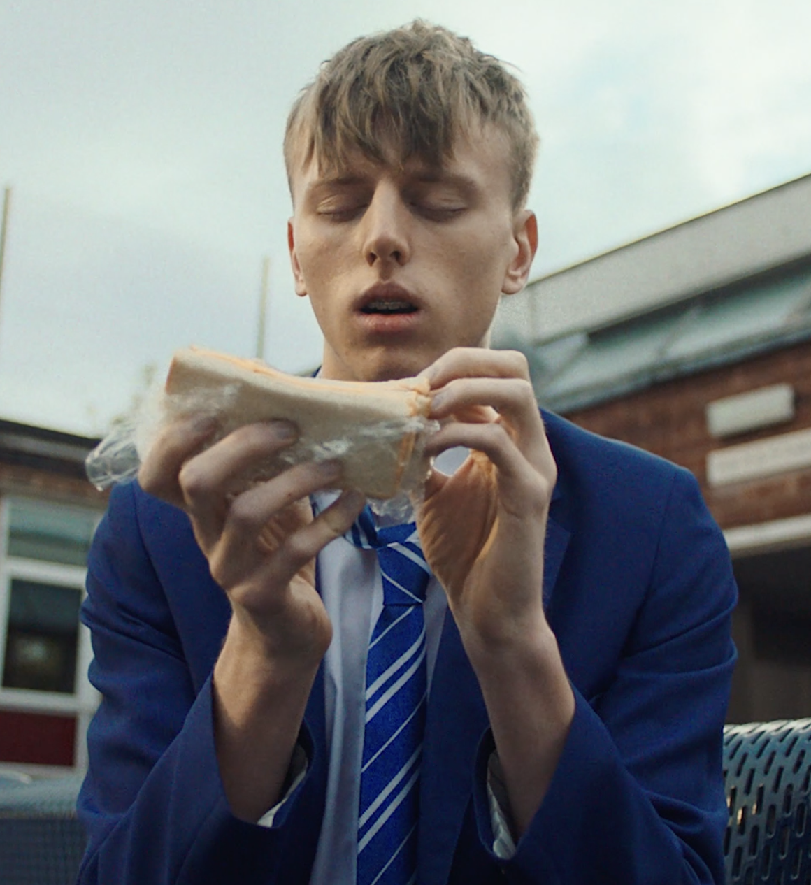 A young man in a blue school uniform and striped tie unwrapping a sandwich, sitting outdoors.