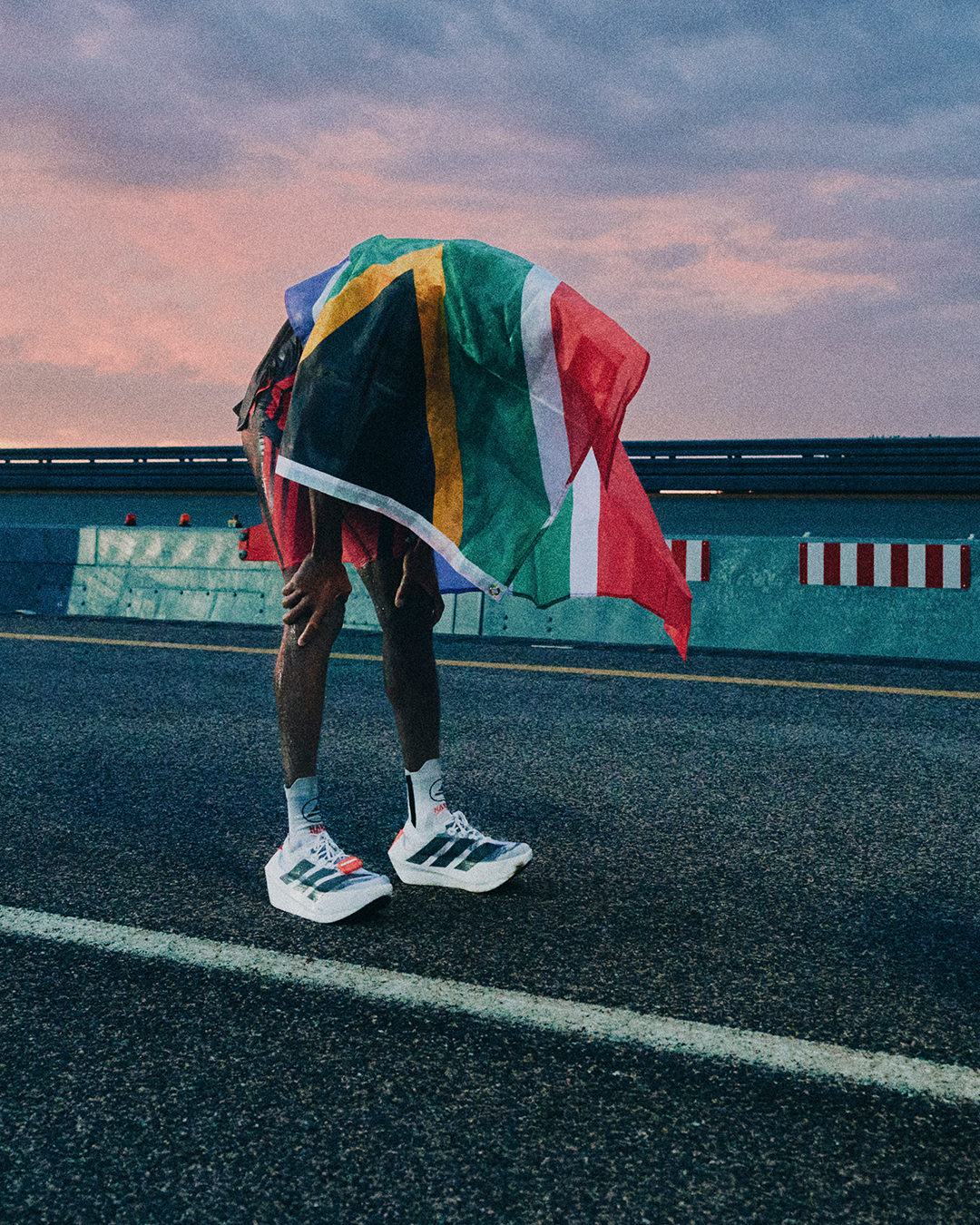 A person standing on a road holding a South African flag covering their face and head, wearing white sneakers with black stripes, at sunset or sunrise with a colorful sky.