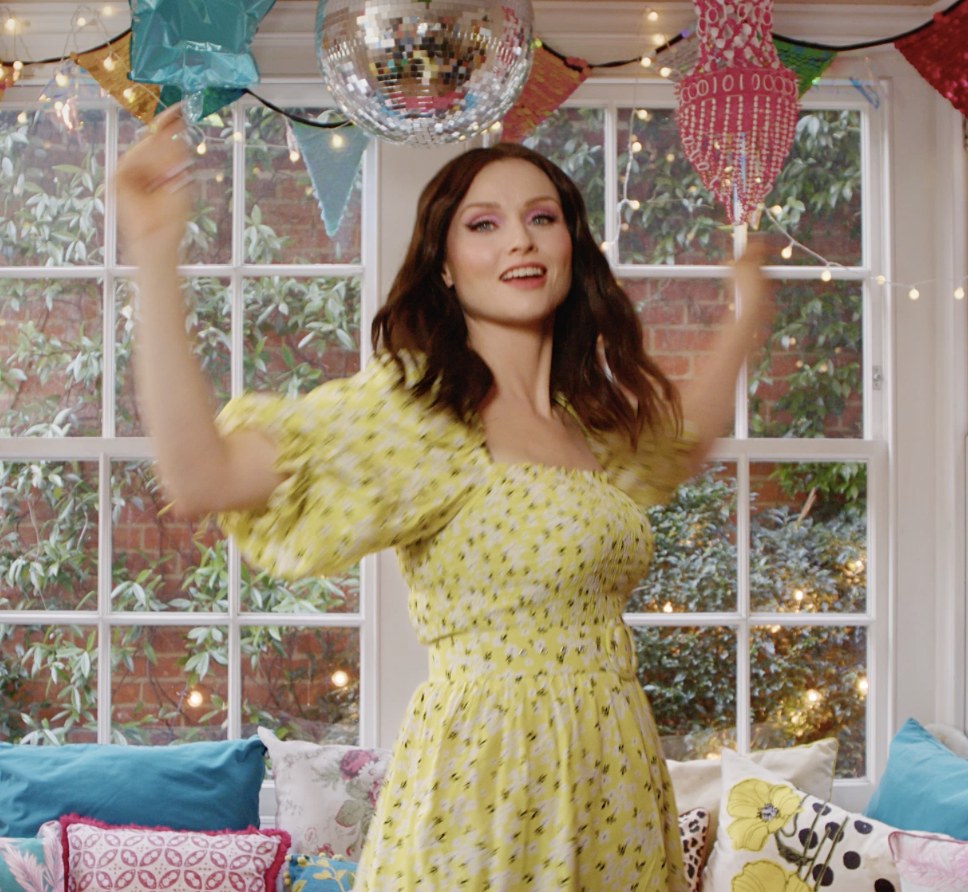 Sophie Ellis Bextor with dark, wavy hair and wearing a yellow dress with small floral print, dancing or posing with her arms raised in a decorated room with colorful paper flags, a disco ball, and cushions on a window seat.