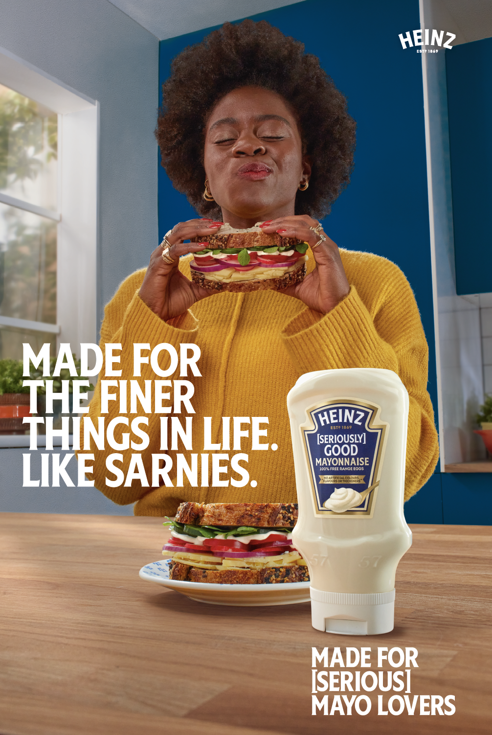 A woman with curly hair in a yellow sweater holding a sandwich with visible layers of vegetables, cheese, and bread, standing in a kitchen. A plate with a similar sandwich and a bottle of Heinz mayonnaise are on the wooden table. Text overlay reads '