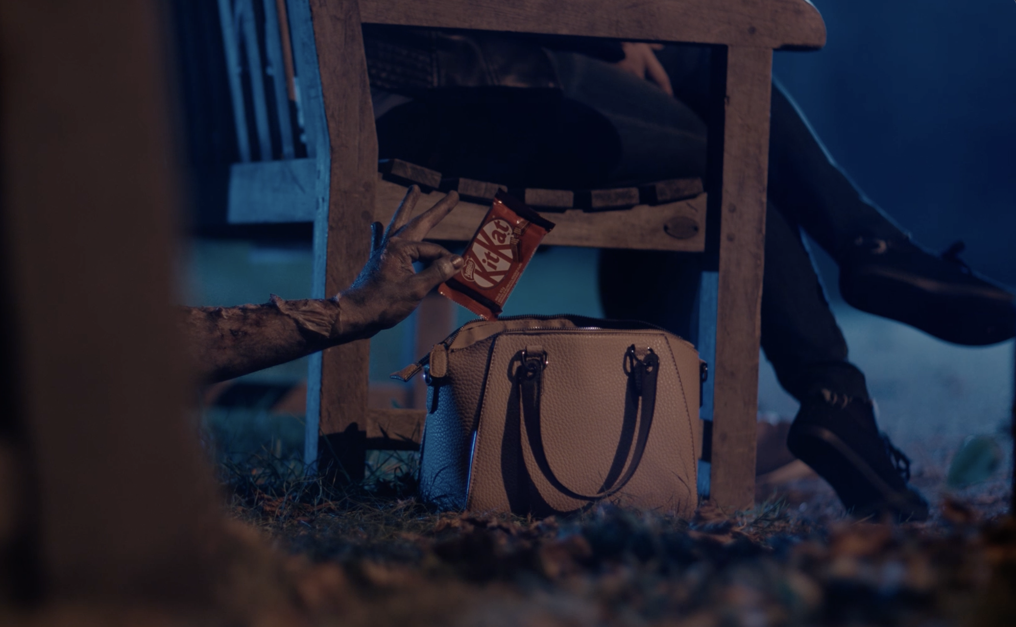 A hand reaches for a Kit Kat chocolate bar on the ground next to a beige handbag beneath a wooden bench. A person is sitting on the bench with legs crossed, wearing dark pants and black shoes, in a dimly lit setting.