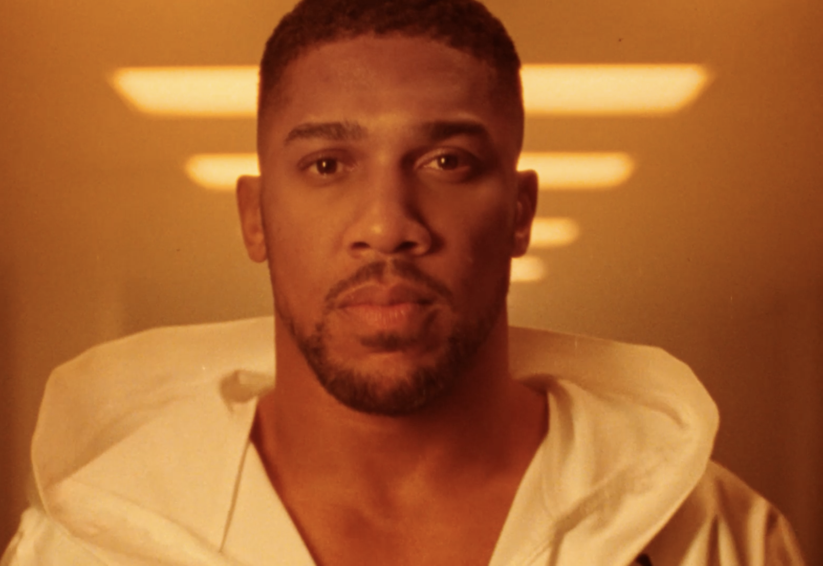 Anthony Joshua wearing a branded Lucozade Alert hoodie, standing against a background with horizontal orange lights.