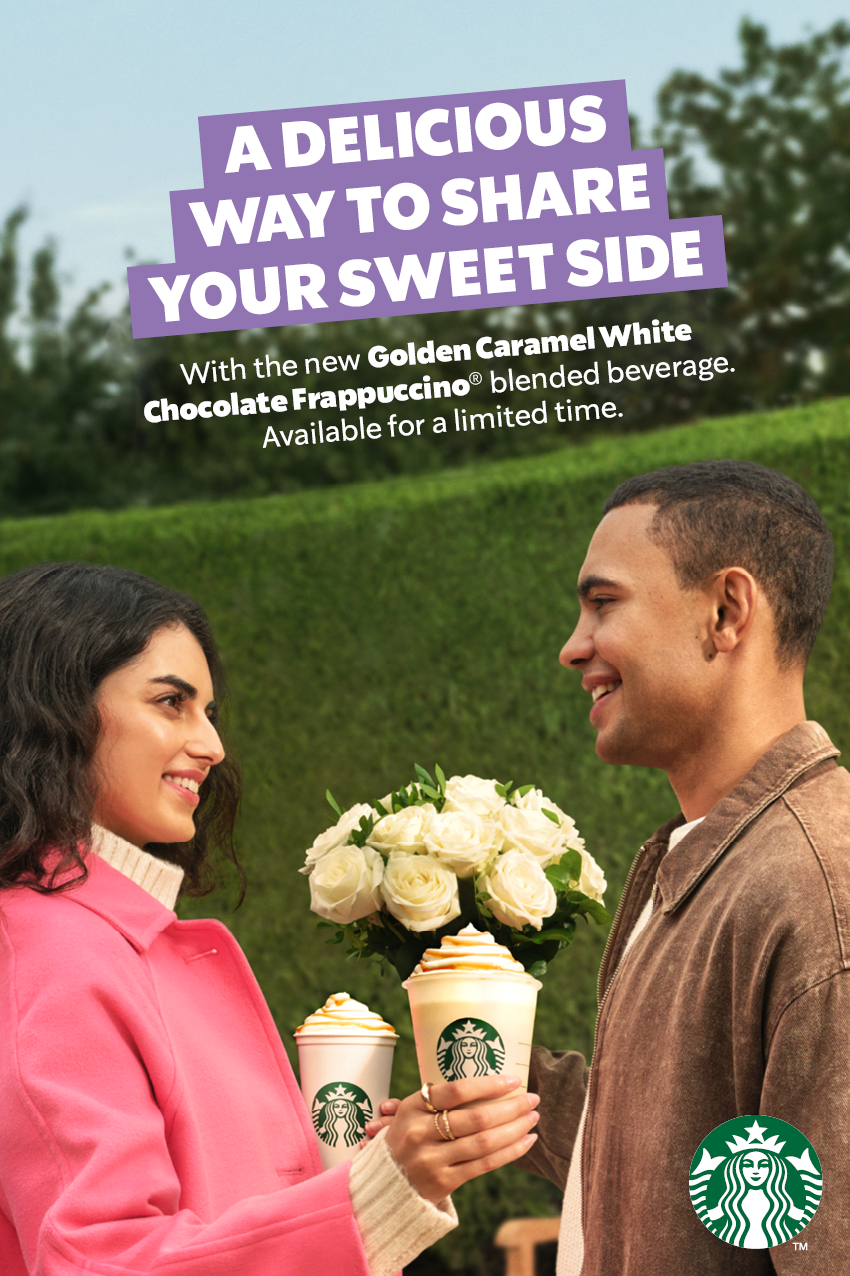 A woman and a man exchanging drinks from Starbucks, holding white cups with green Starbucks logos. The woman is wearing a pink coat, and the man is in a brown jacket, smiling at each other in a park setting with greenery and trees in the background.