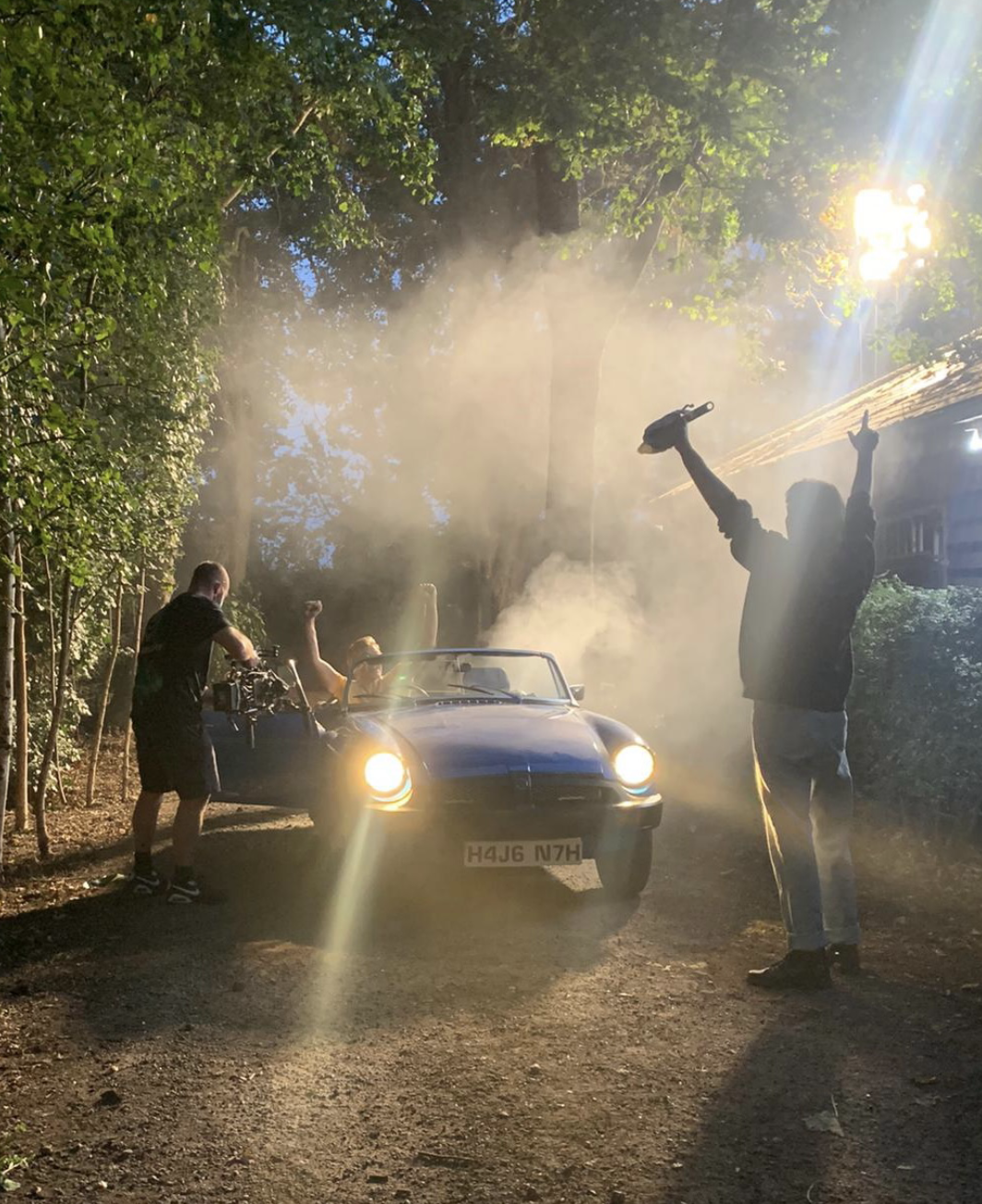 People filming a vintage car surrounded by trees and smoke during sunset or sunrise, with one person celebrating.