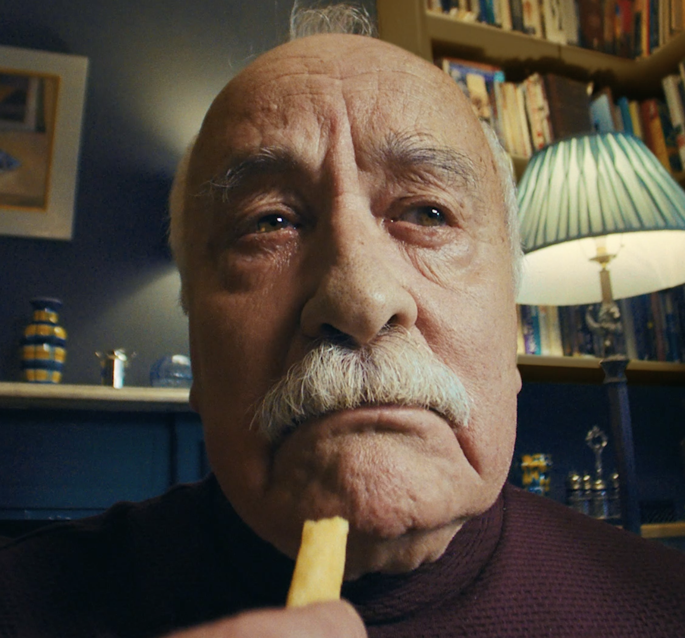 Close-up of an elderly man with a serious expression holding a French fry near his mouth, in a room with a bookshelf, lamp, and decorative ceramics in the background.