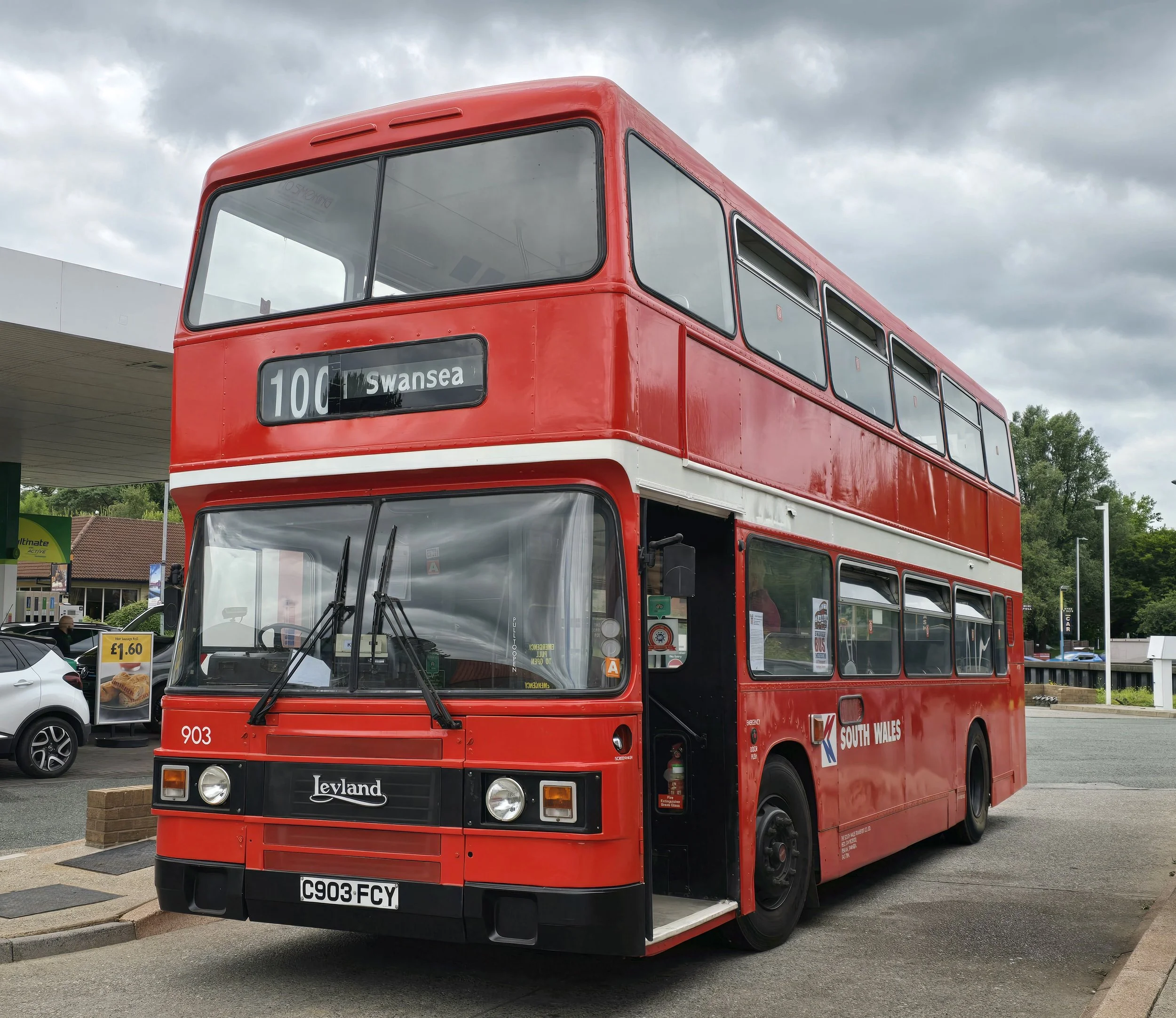 A red double-decker bus with a white stripe, labeled South Wales, parked at a gas station on a cloudy day. The bus displays route 100 to Swansea.