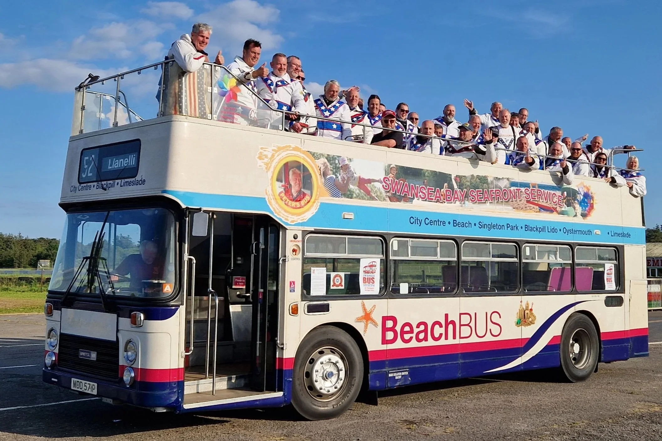 A double-decker sightseeing bus labeled 'BeachBUS' with a group of people on the upper deck, many giving thumbs up and waving. The bus has signage indicating it serves the Swansea Bay Seafront, City Centre, Beach, Singleton Park, Blackpill Lido, Oystermouth, and Limeslade areas. The backdrop features a blue sky and some greenery.