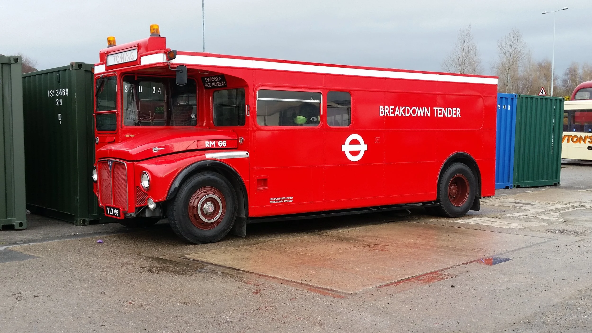 Vintage red bus with white markings, labeled 'BREAKDOWN TENDER' and 'Swansea Bus Museum,' parked on a lot next to a green shipping container and another bus in the background.
