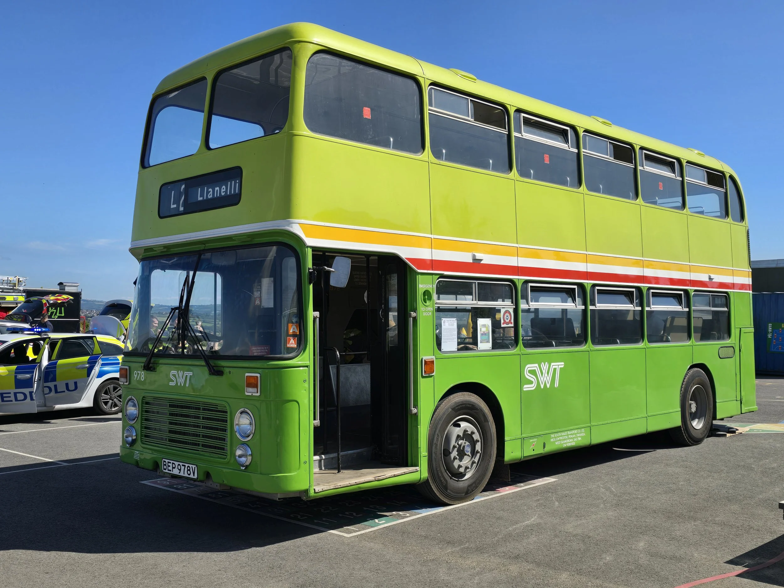 A bright green double-decker bus parked in a lot with police cars nearby on a clear, sunny day.