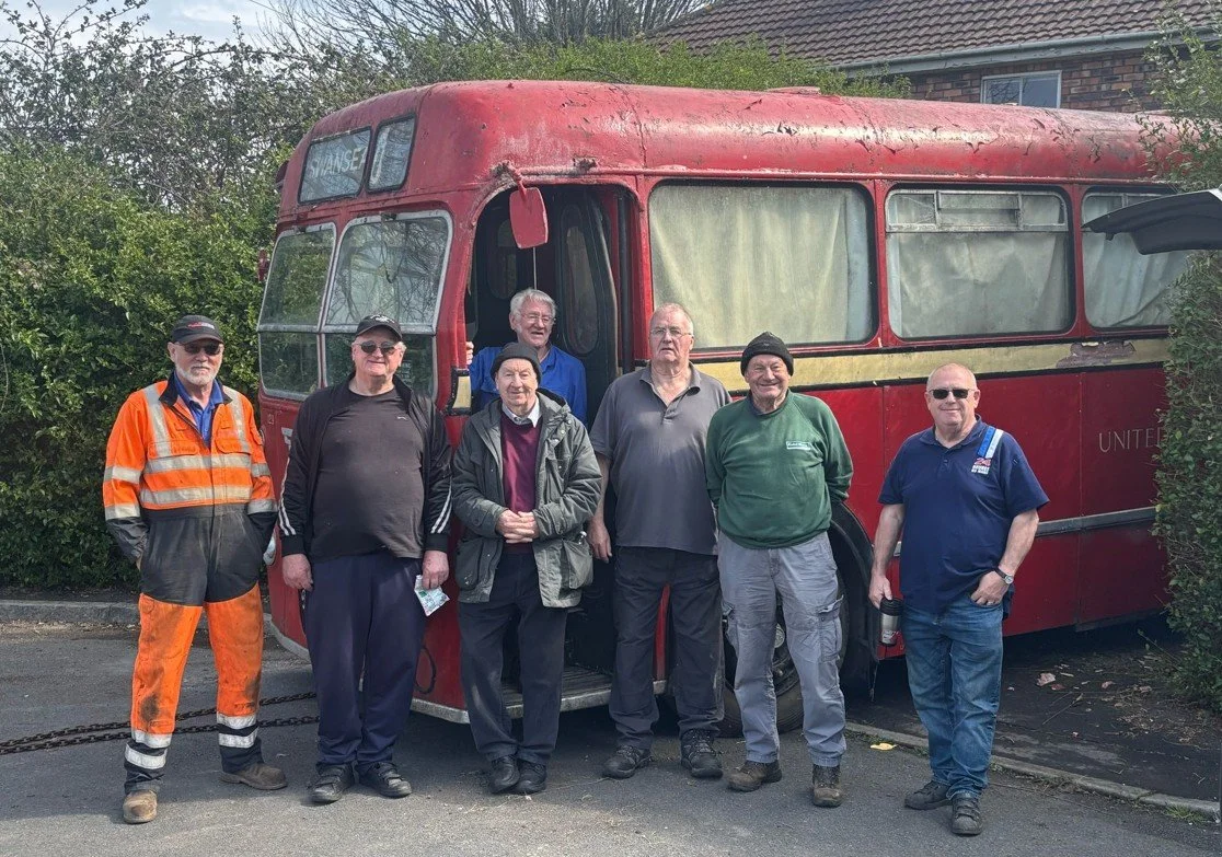A group of seven men standing in front of a vintage red double-decker bus. The men are dressed casually, with one in an orange reflective work jacket and pants, and others in jackets, sweaters, and hats. The bus appears old and partly weathered, surrounded by greenery.