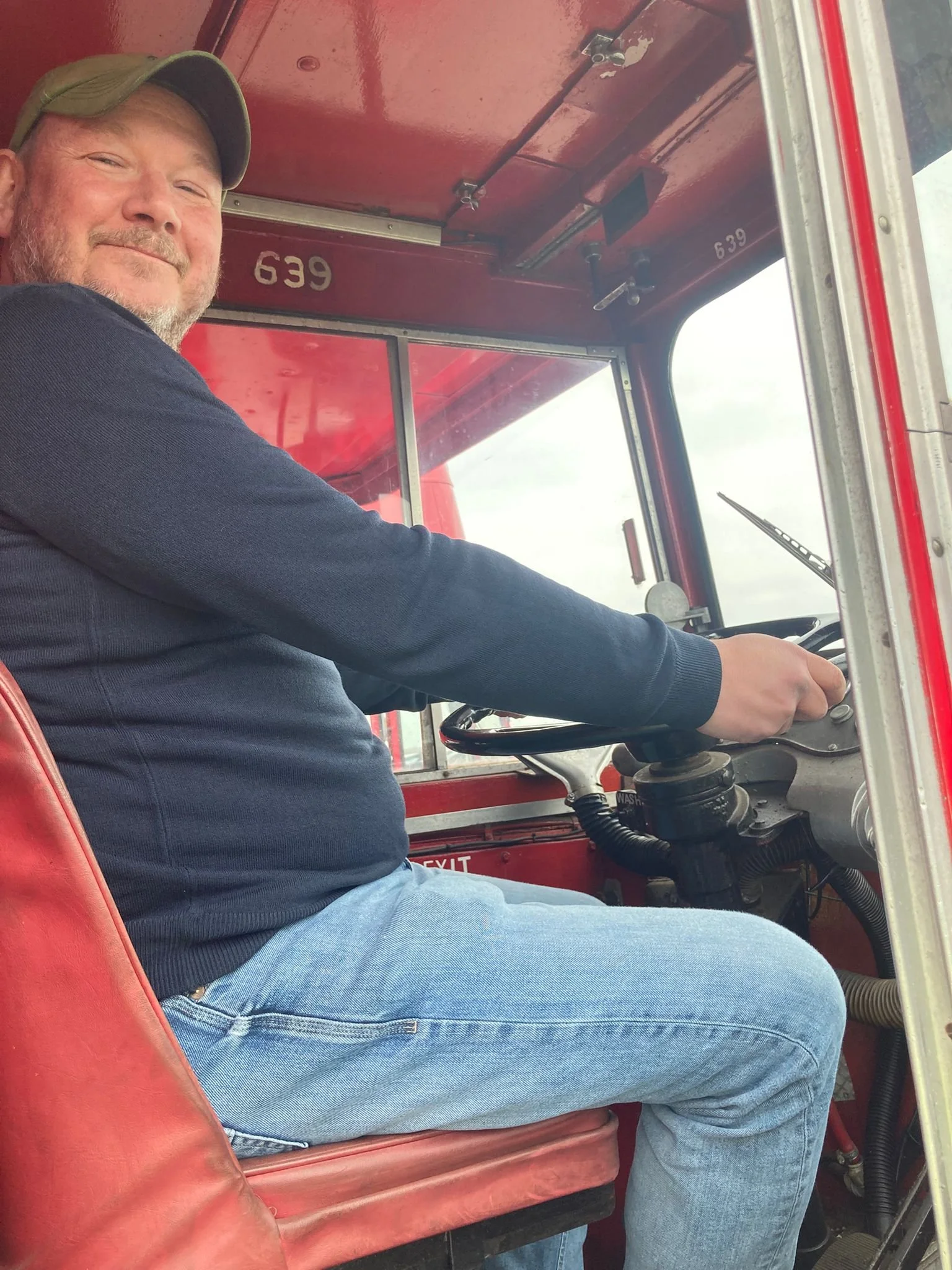 A man sitting in the driver's seat of a fire truck, smiling, with a steering wheel and dashboard in view.