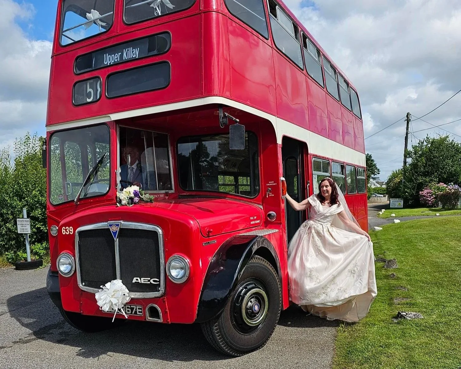 Woman in wedding dress standing on the ladder of a vintage red double-decker bus, holding the side of the bus with a smile, in an outdoor setting with green grass and trees.
