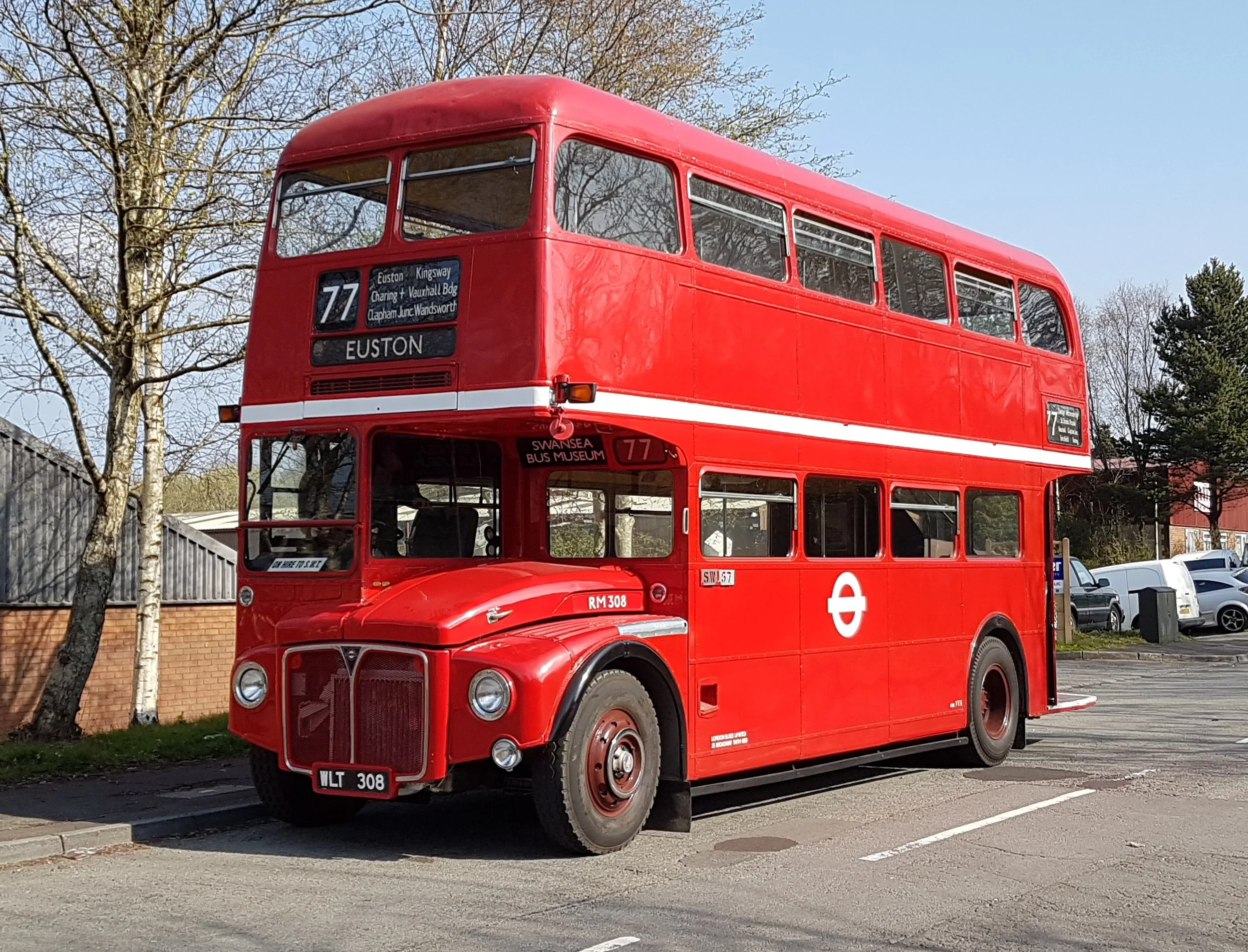 Red double-decker bus parked in a lot with trees and cars in the background.