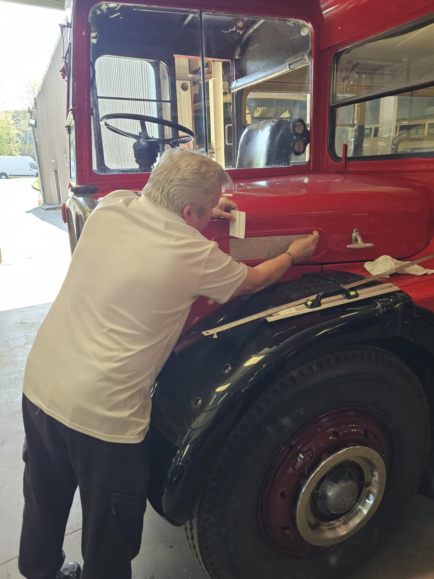 Woman in wedding dress standing on the ladder of a vintage red double-decker bus, holding the side of the bus with a smile, in an outdoor setting with green grass and trees.