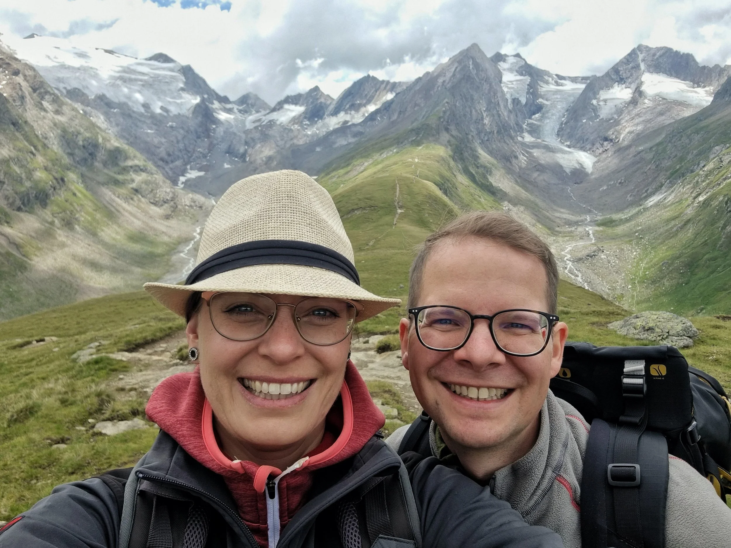 Zwei lächelnde Menschen, eine Frau und ein Mann, in den Bergen mit grünen Hängen, Gipfeln und Schneefeldern im Hintergrund. Sie tragen Wanderausrüstung.