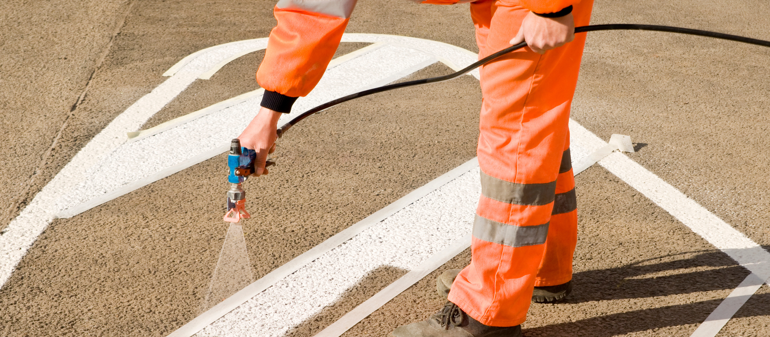 Worker in orange safety clothing applying white paint to asphalt road using spray equipment, creating a white line.