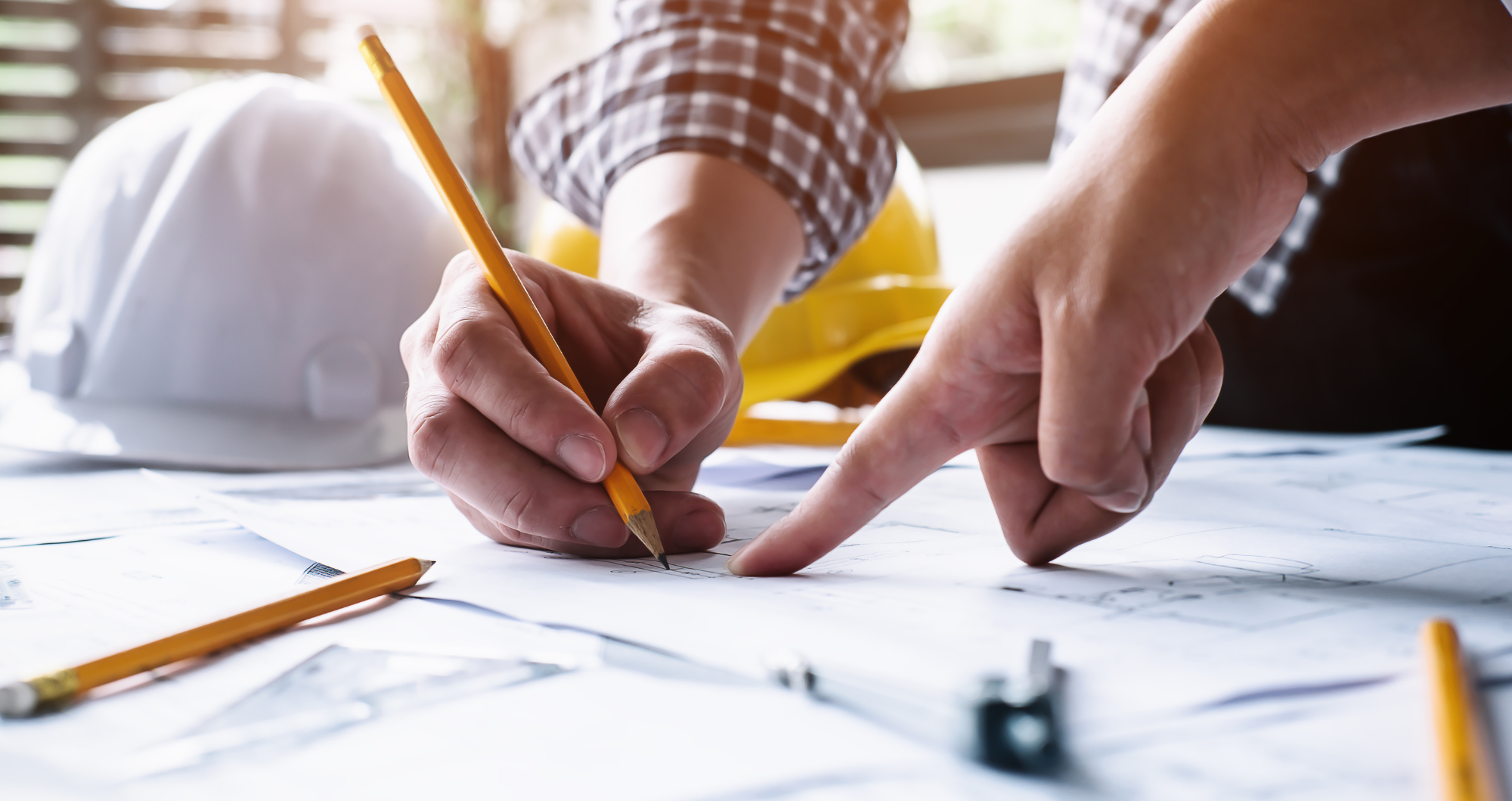Person in a plaid shirt pointing and drawing on blueprints with a pencil, a white construction helmet and a yellow hard hat on the table.