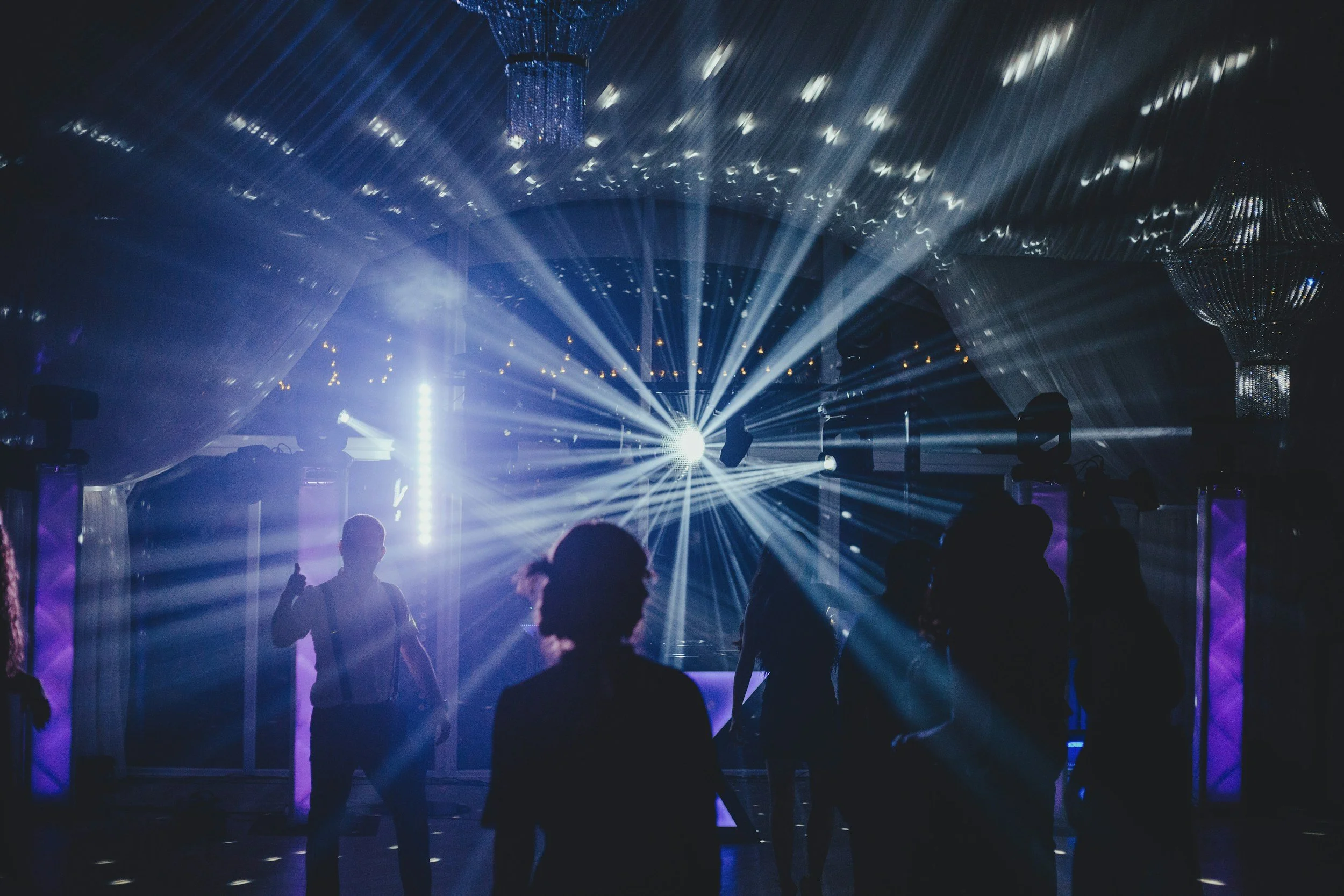 People dancing at a nightclub with bright blue and white lights and large chandeliers.