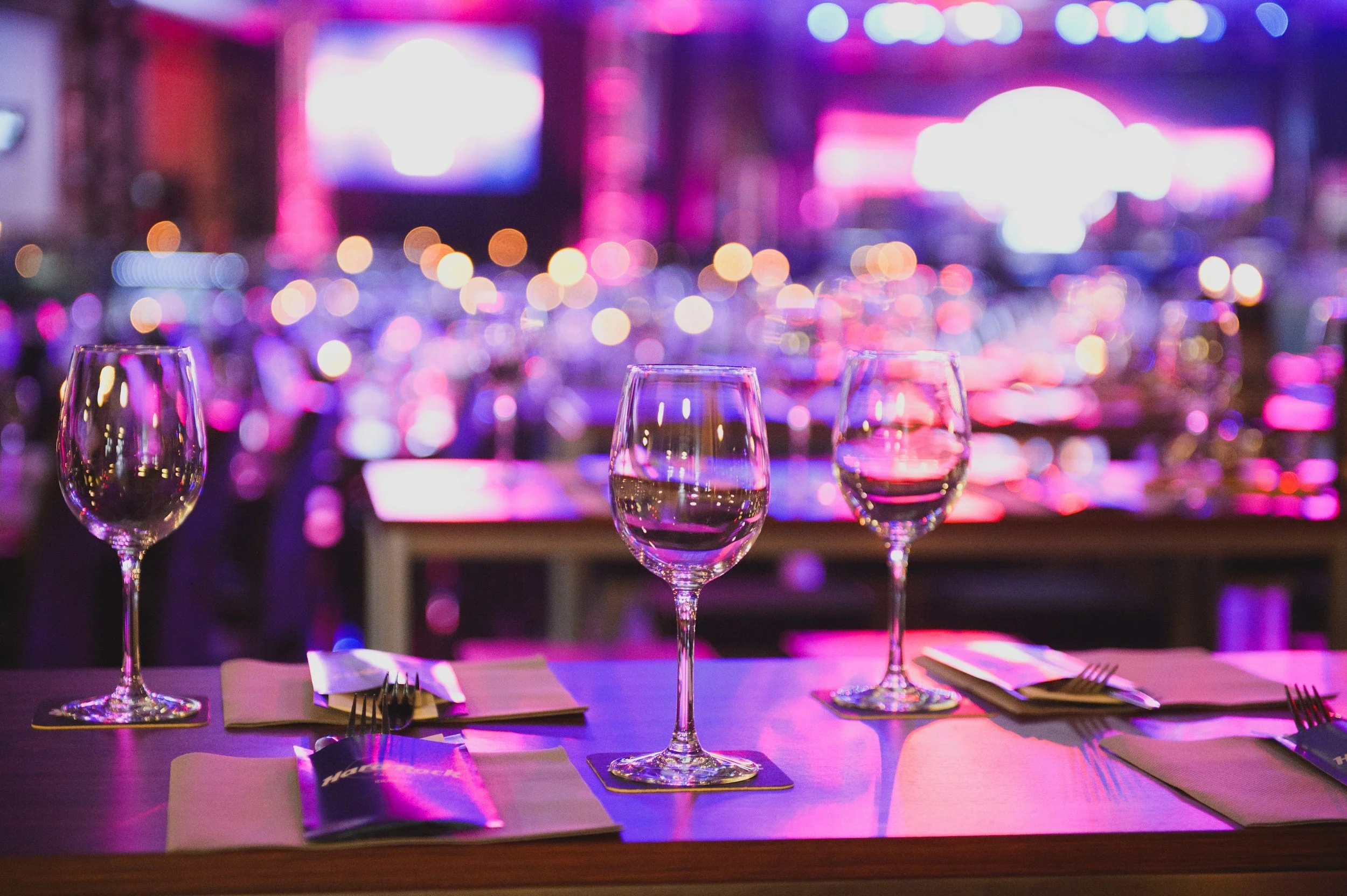 Three empty wine glasses on a table with white napkins and cutlery in a dimly lit setting with pink and purple neon lights, suggesting a nightlife or bar scene.