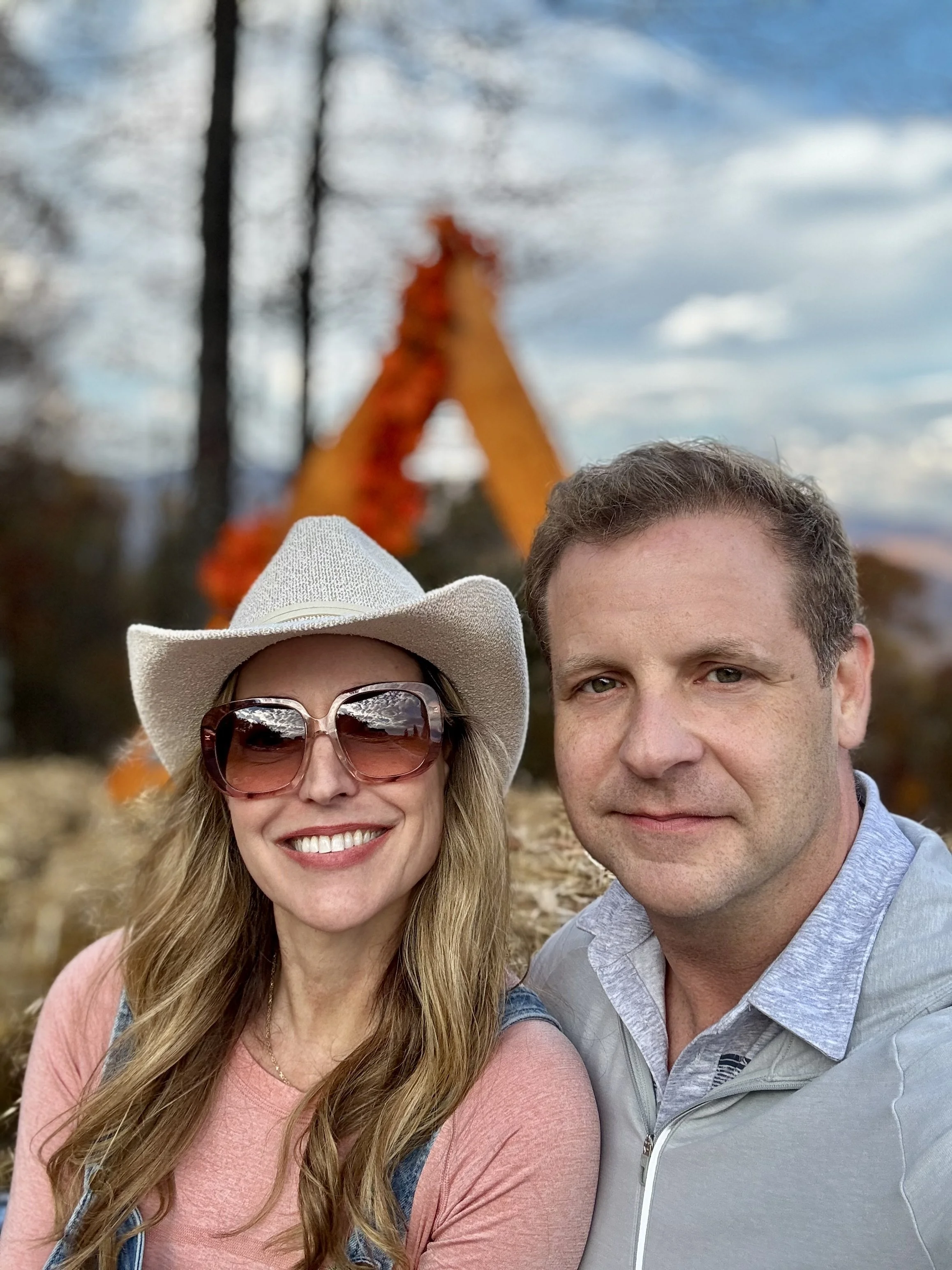 A smiling woman wearing large sunglasses and a cowboy hat, and a man with short hair, both outdoors in front of a blurred autumn scene with trees and a decorative orange arch in the background.