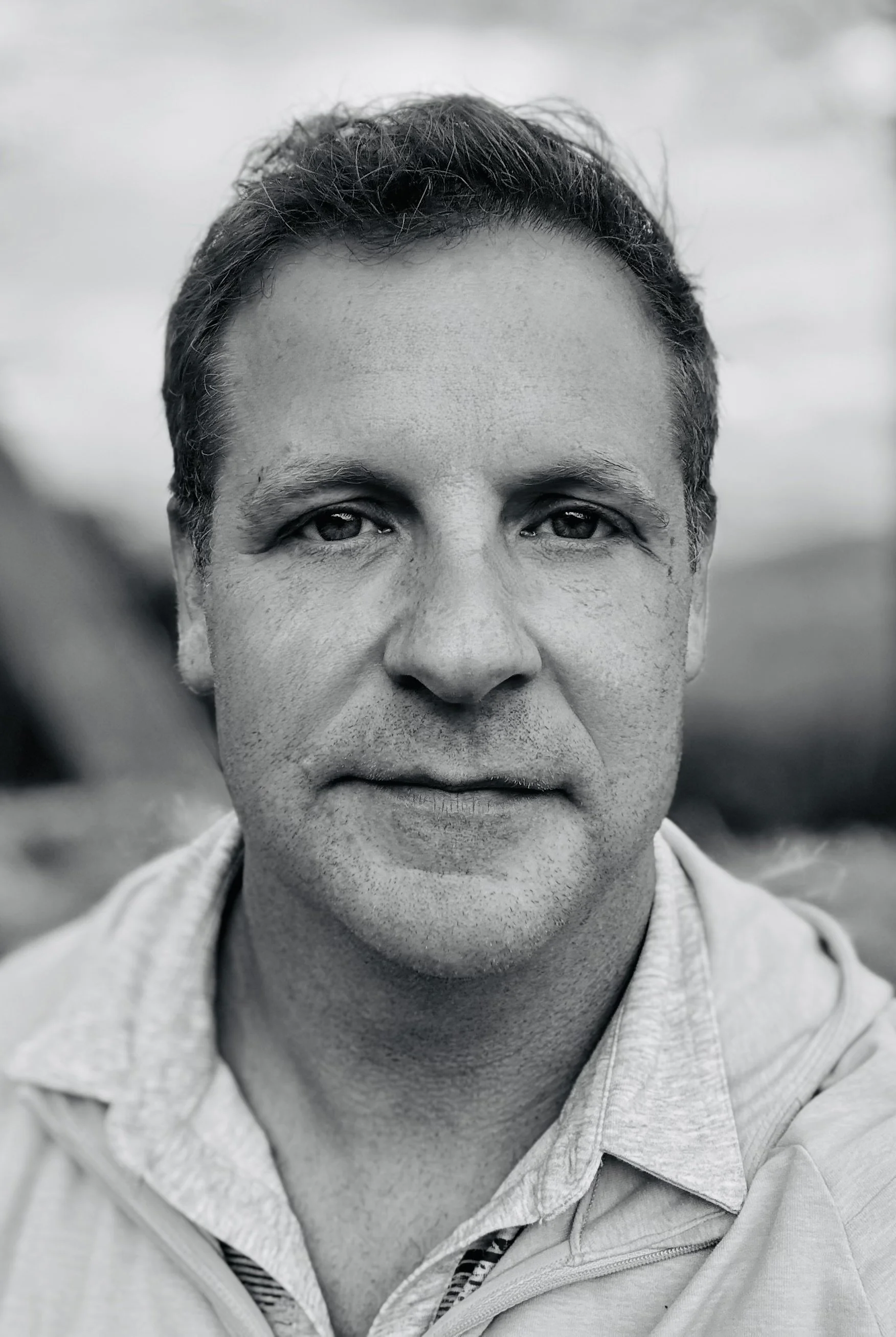 Close-up black and white portrait of a middle-aged man with short, dark hair, wearing a collared shirt and jacket, outdoors with a blurred landscape in the background.