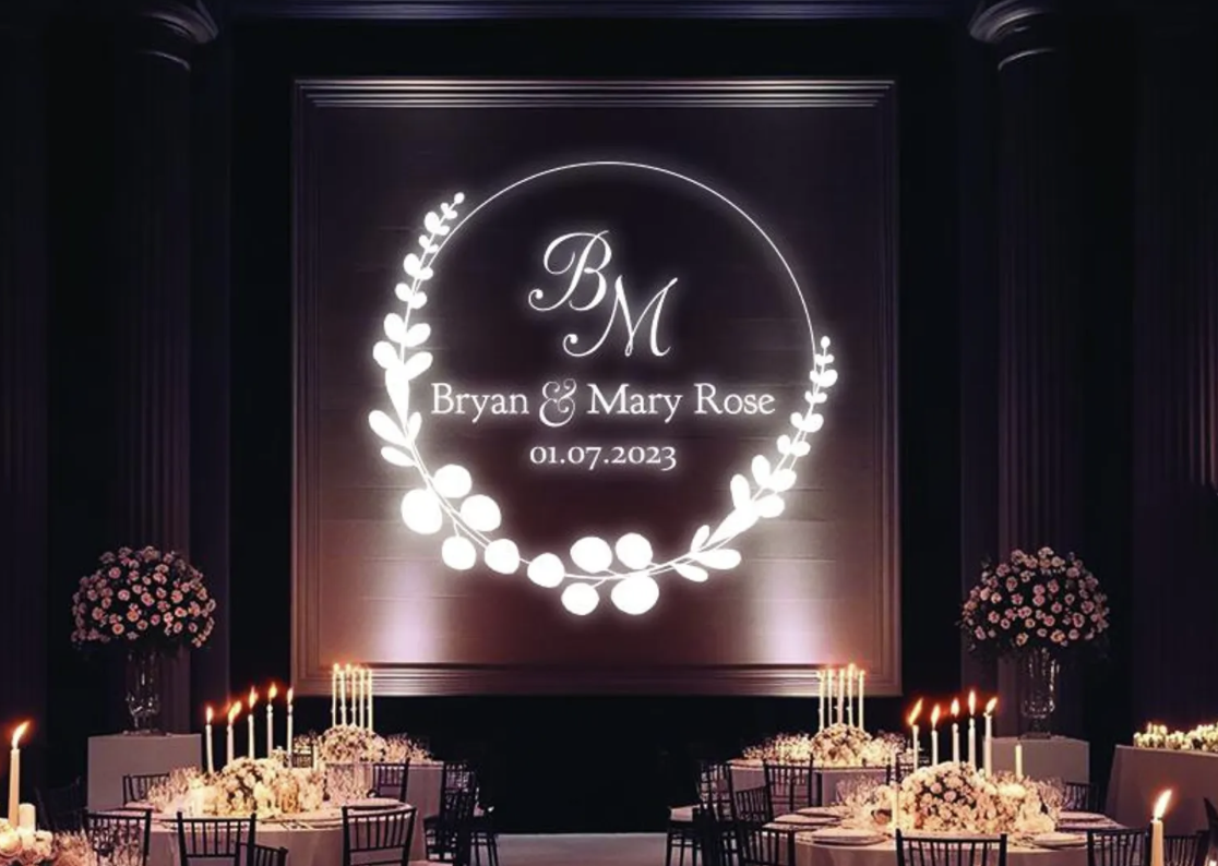 Wedding reception with floral arrangements and candlelit tables, projected sign with initials 'BM', names 'Bryan & Mary Rose', and date '01.07.2023'.