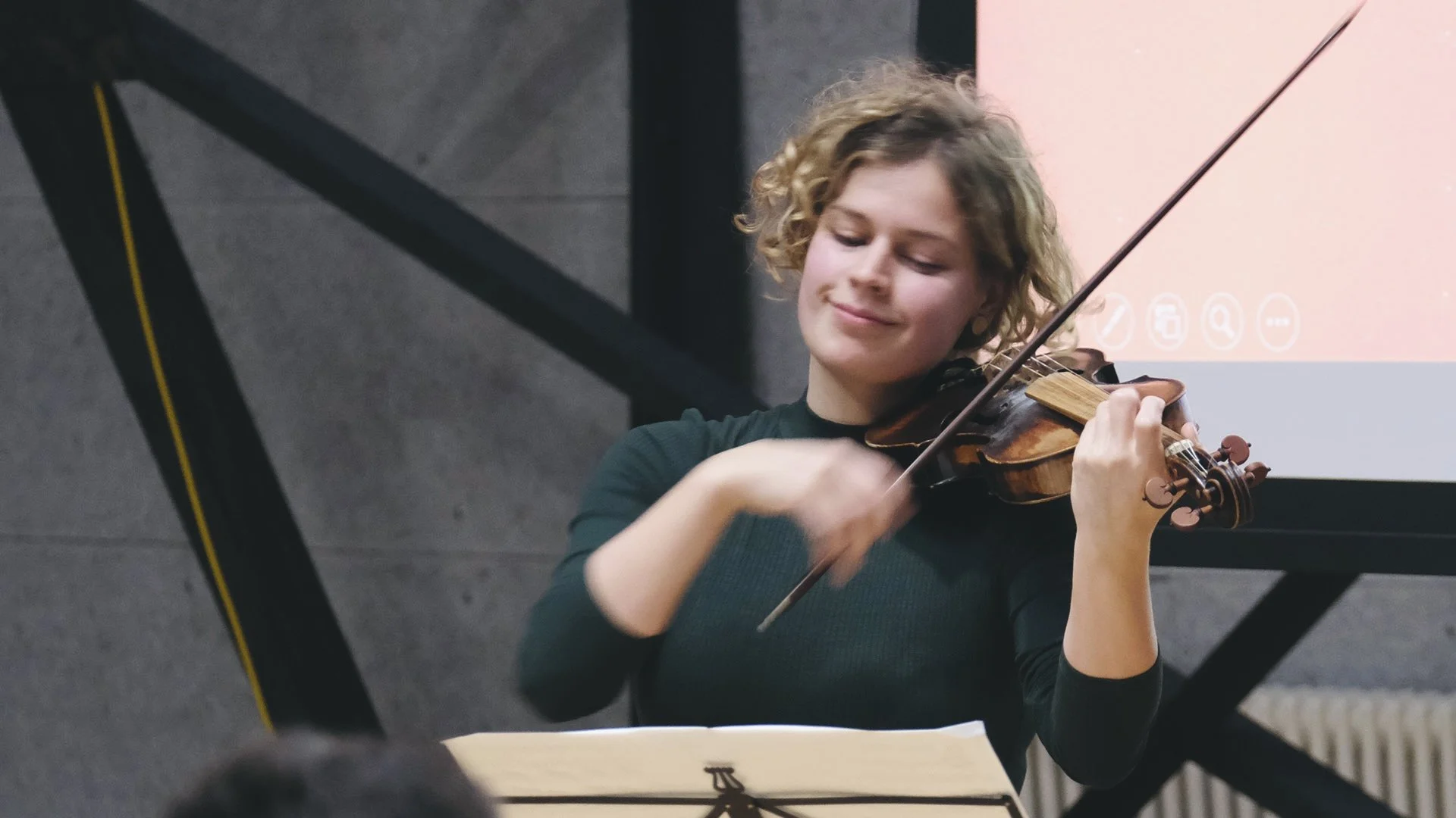 A young woman with curly blonde hair playing the violin during a performance or practice session, with a music stand and screen in the background.
