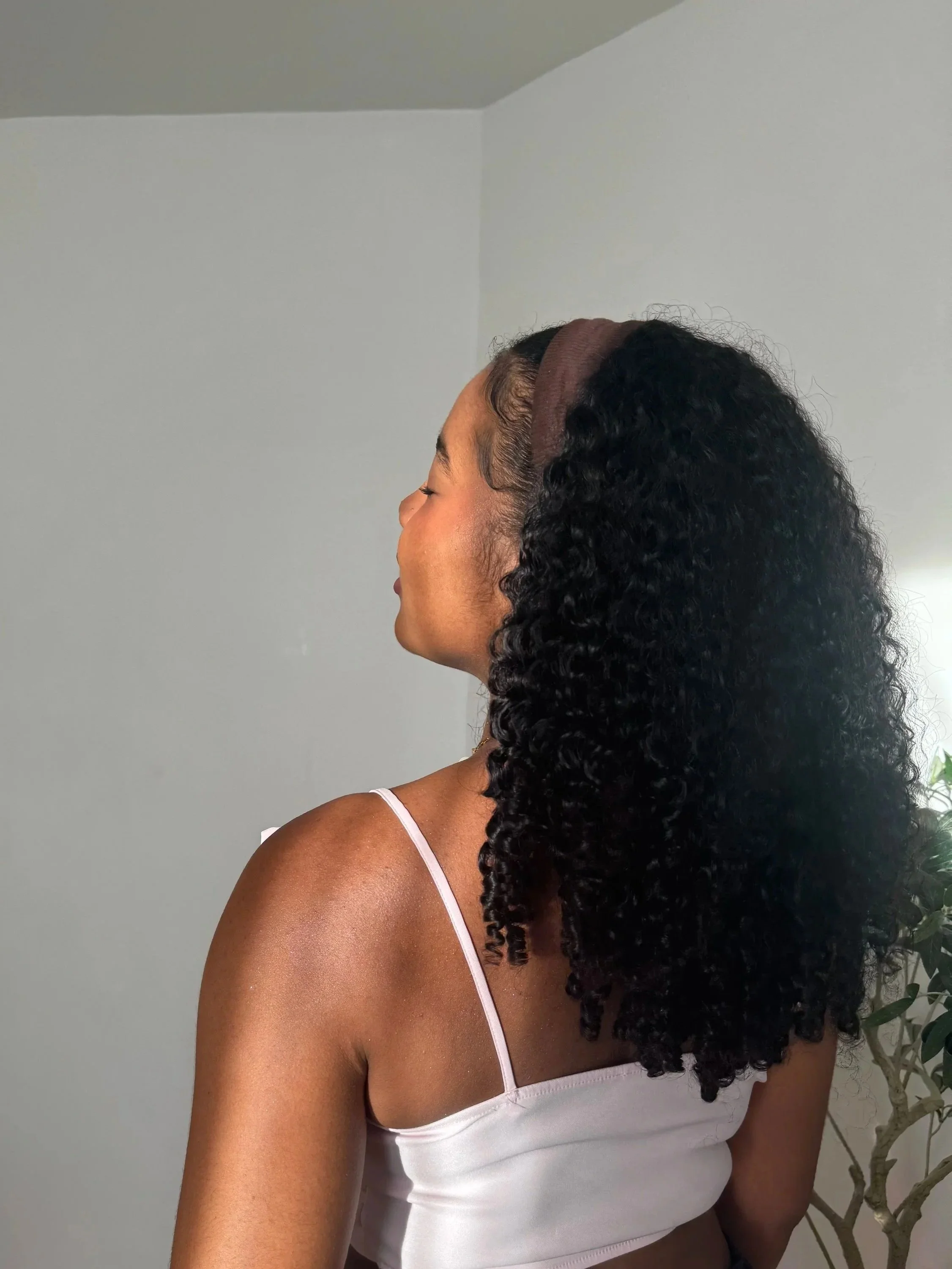 Une femme vue de profil avec des cheveux bouclés noirs, portant un bandeau marron et un haut blanc à fines bretelles, se tenant près d'une plante dans une pièce avec un mur blanc.