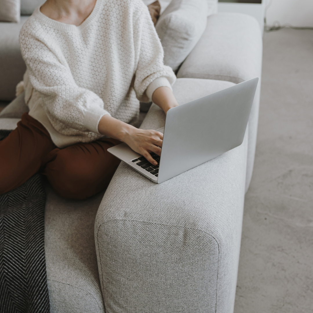 Person sitting on a light-colored sofa using a silver laptop, wearing a white knitted sweater and brown pants.